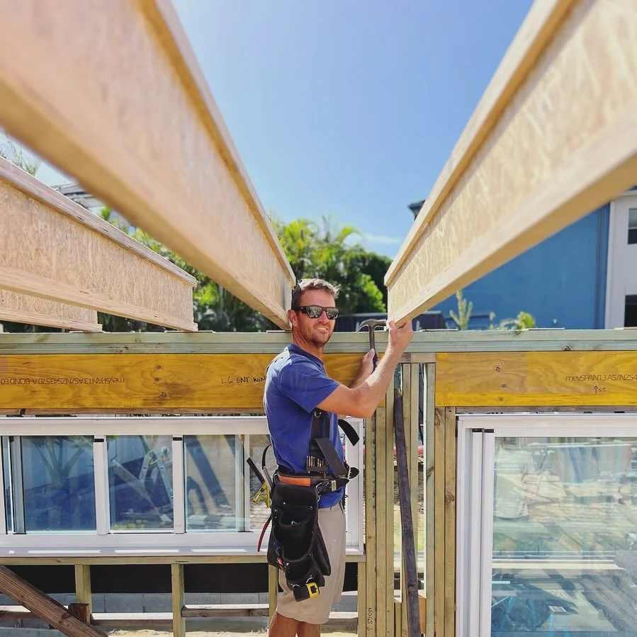 A construction worker wearing sunglasses and a tool belt is standing inside a building under construction, holding a long wooden beam, with a clear blue sky and green trees in the background.