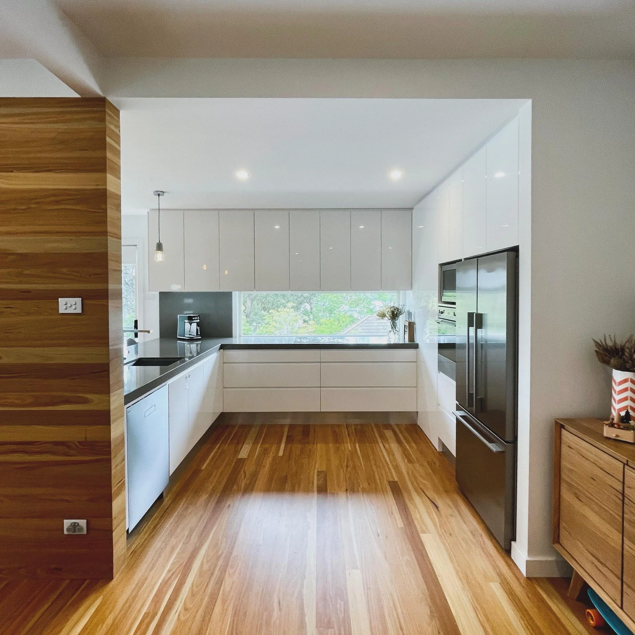 Home renovation featuring a modern kitchen with white cabinets, grey concrete countertops, stainless steel appliances, hardwood flooring, a feature timber wall, and a large window overlooking greenery.