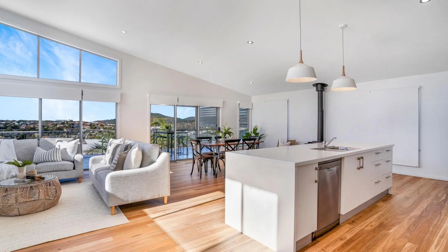 Open-concept living room and kitchen with large windows, white walls, wooden floors, and modern furniture, including a white sofa, round wicker coffee table, dining table with chairs, and pendant lights.