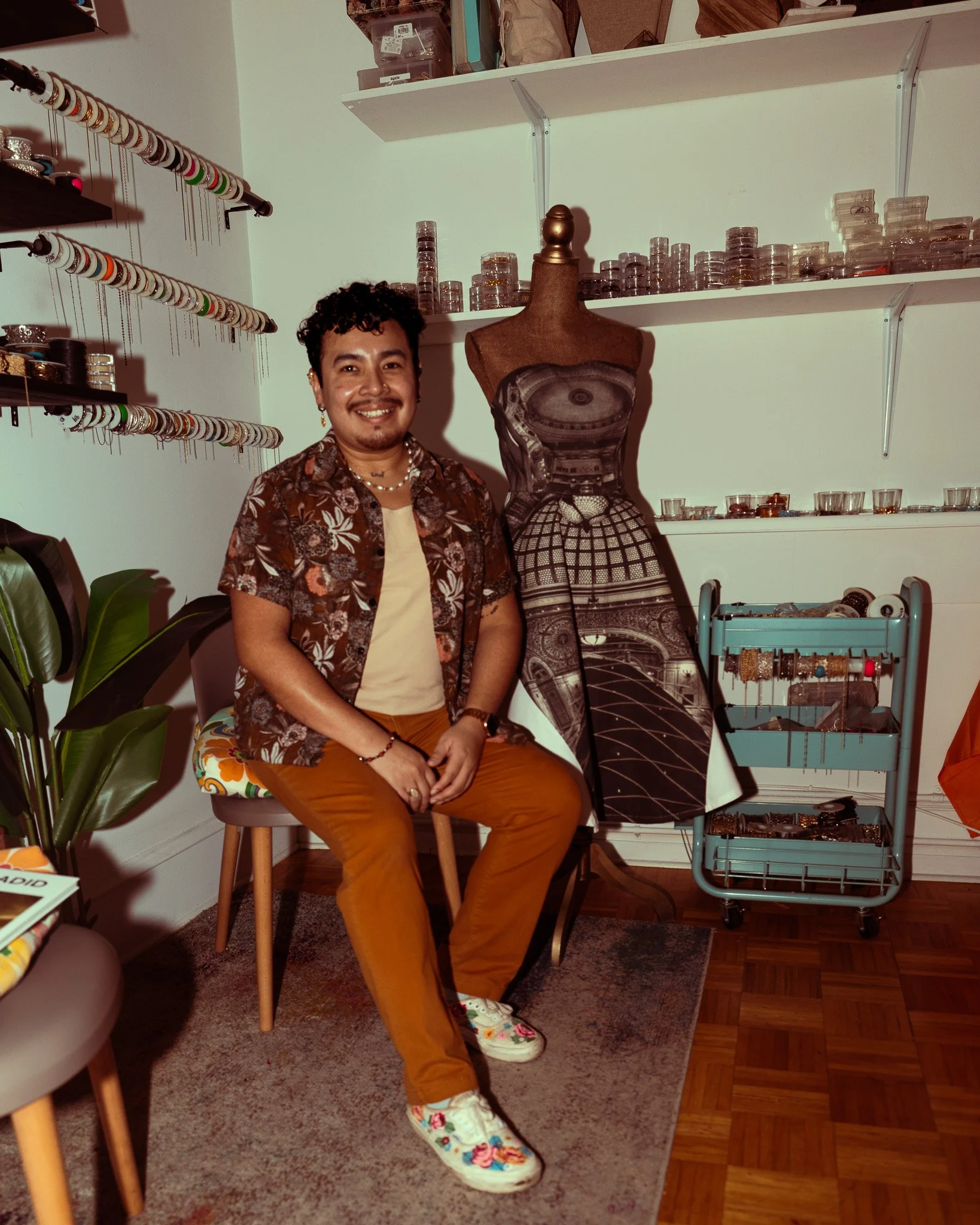 A person sitting on a chair in a room with jewelry displays, mannequins, and shelves filled with jewelry supplies and tools.