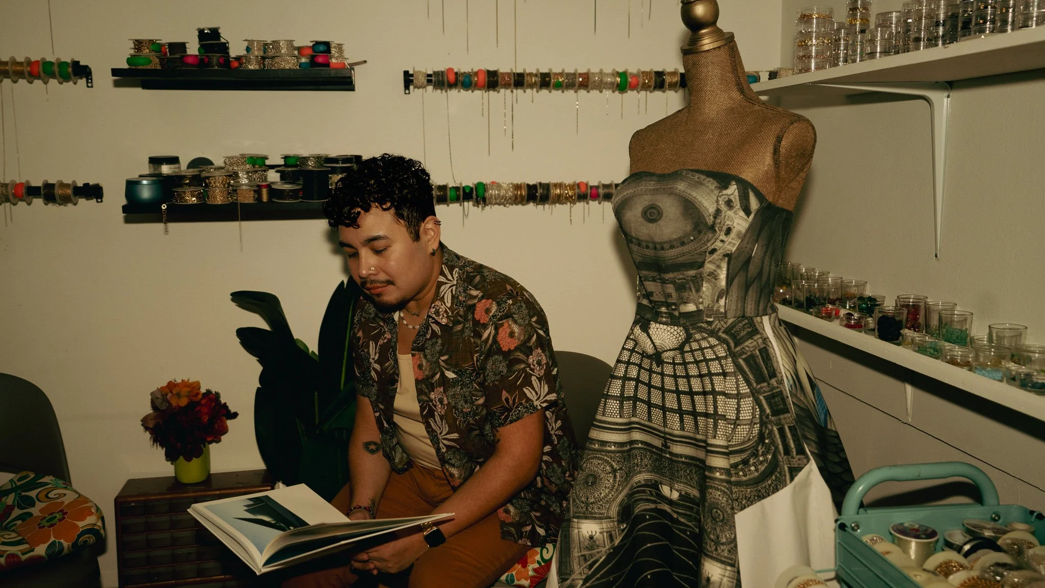 A person sitting on a chair in a room decorated with shelves of jewelry and watch collections, holding and looking at an open photo album. There is a dress form with a dress, a vase with flowers, and a cart with accessories nearby.