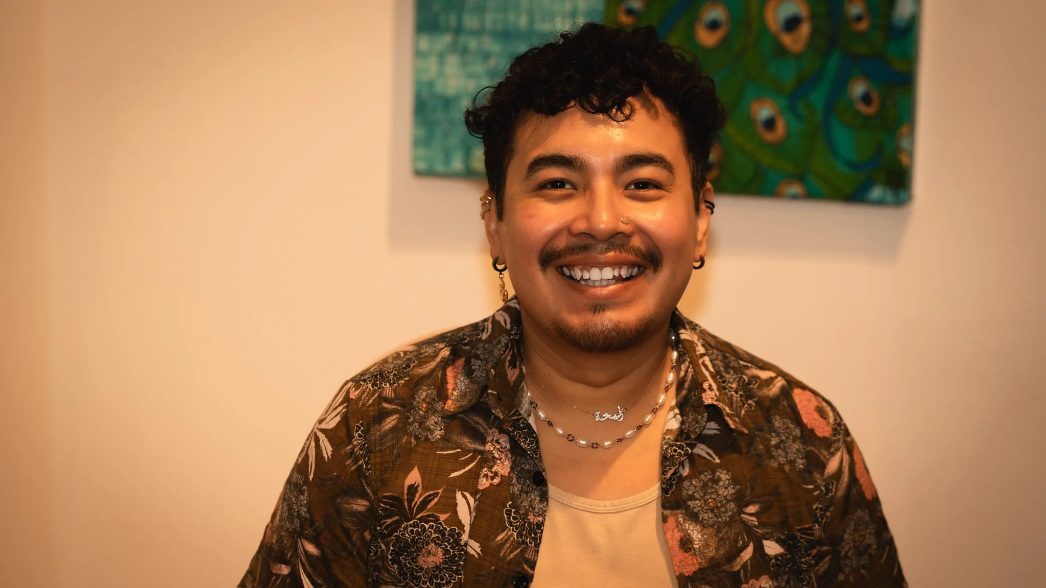 A smiling young man with dark, curly hair and a mustache, wearing a floral patterned shirt, layered necklaces, and earrings, standing indoors with a colorful abstract painting in the background.