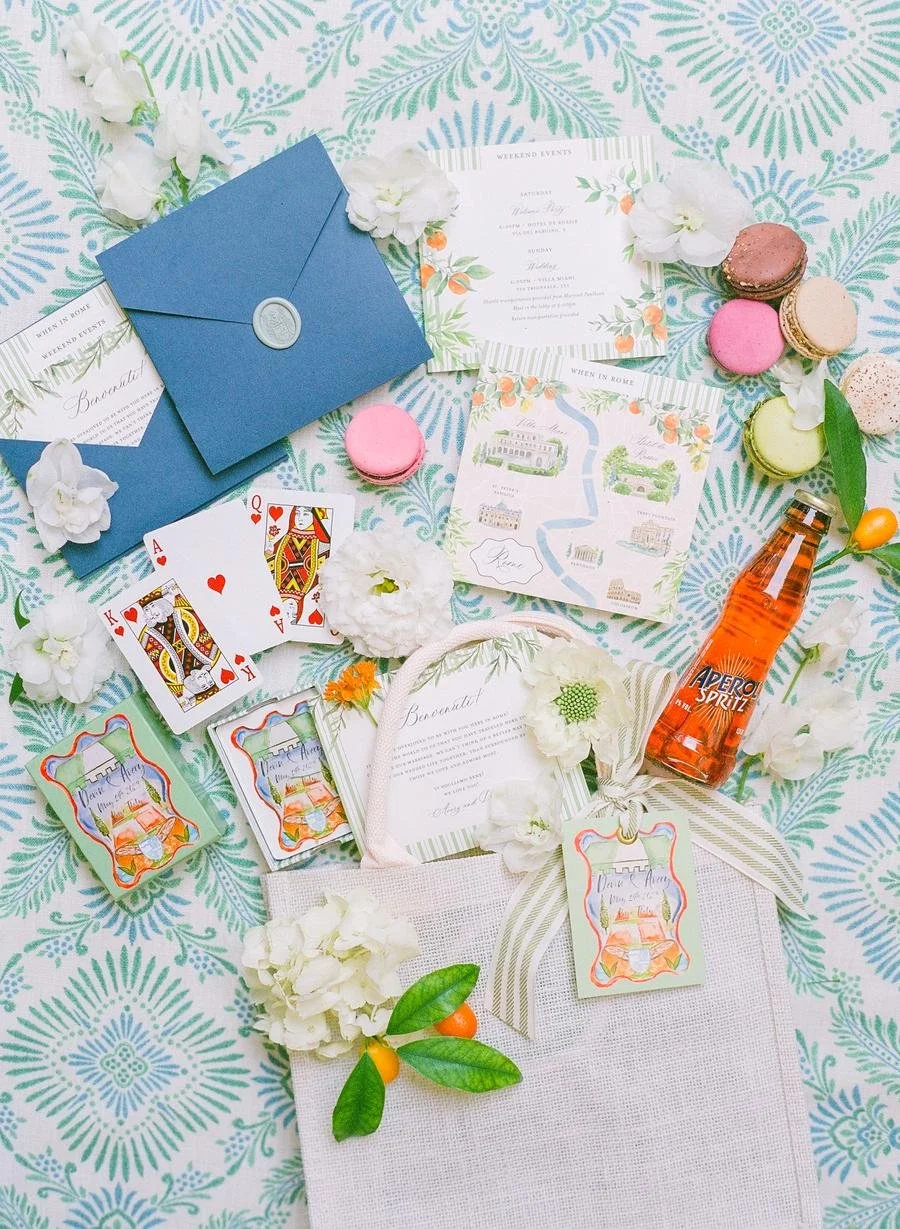 Flat lay of wedding invitations and stationery on a patterned background with blue envelopes, playing cards, colorful macarons, white flowers, and an Aperol Spritz bottle.
