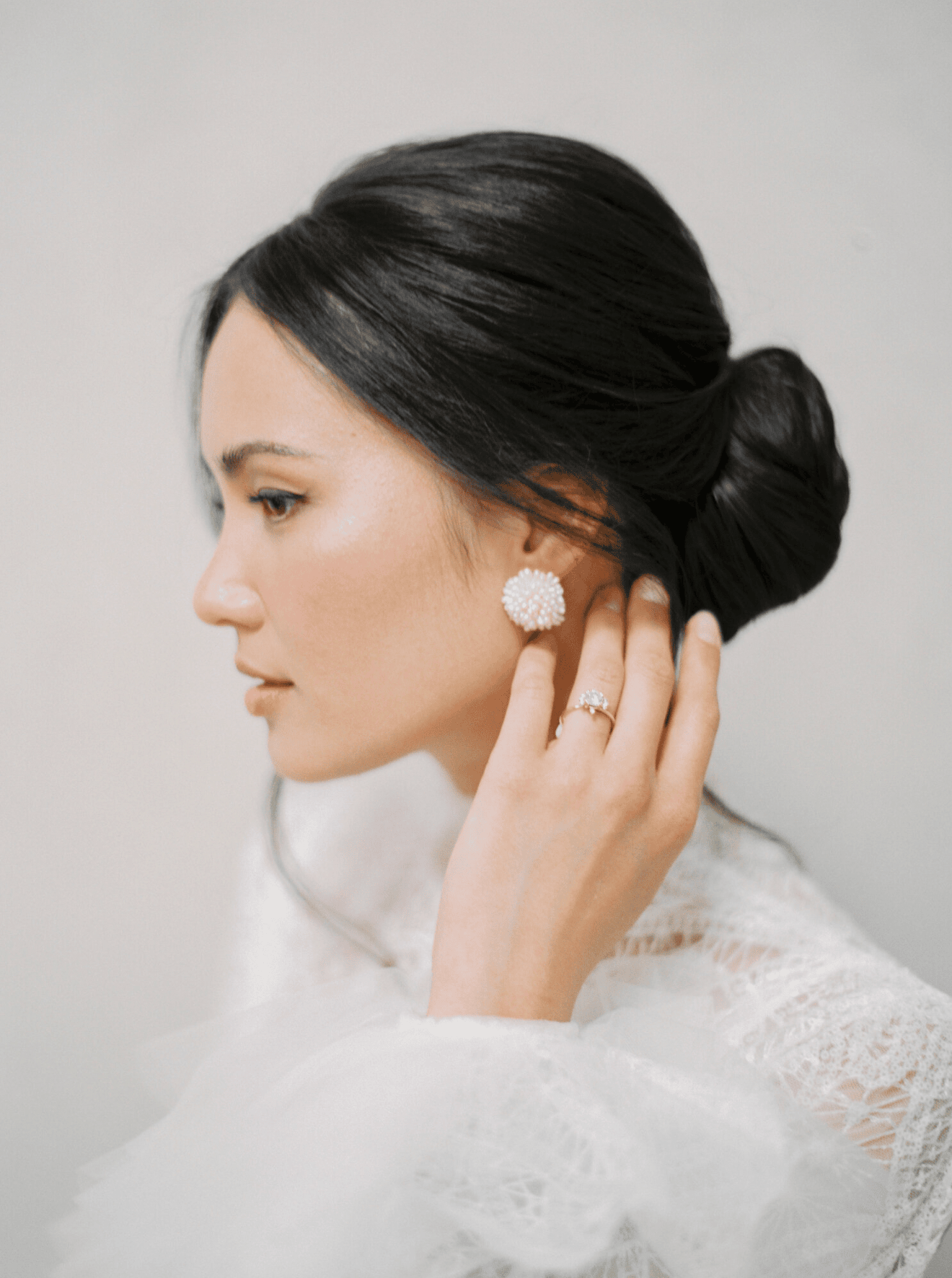 Elegant woman in white lace attire, with a pearl cluster earring, her hand touching her ear, and dark hair styled in a bun.