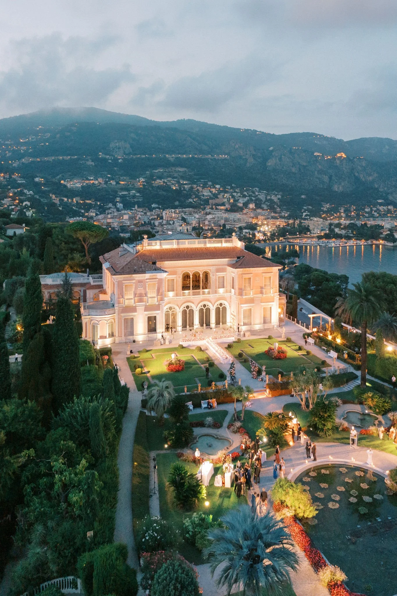 Aerial view of a grand villa illuminated at dusk, surrounded by lush gardens and overlooking a coastal town and hills.