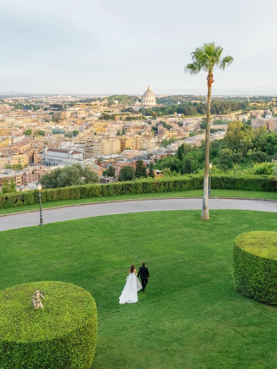 A couple in formal attire walking on a lush green lawn with a cityscape view in the background, featuring a prominent dome. The scene includes a palm tree and trimmed shrubs.