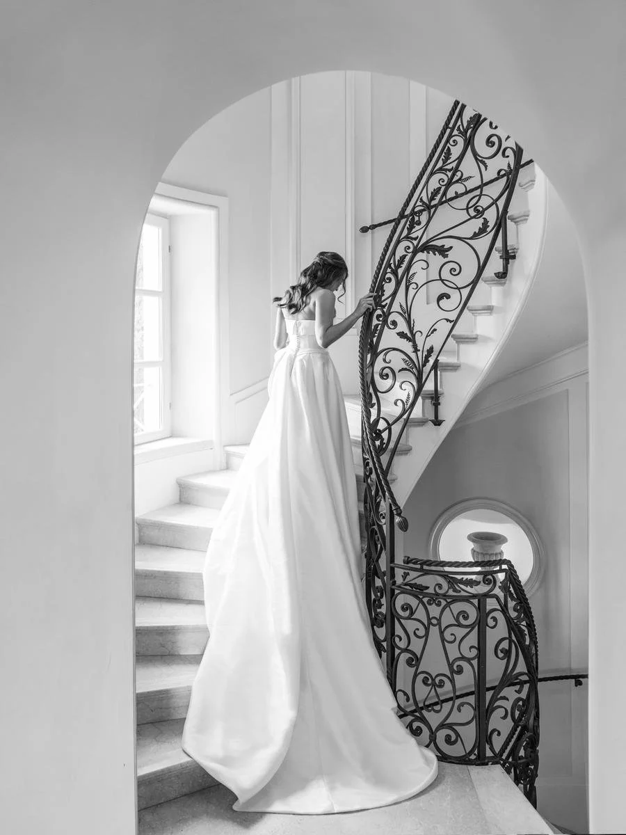 Bride in a flowing wedding dress ascending a spiral staircase with ornate black railings in a brightly lit room.