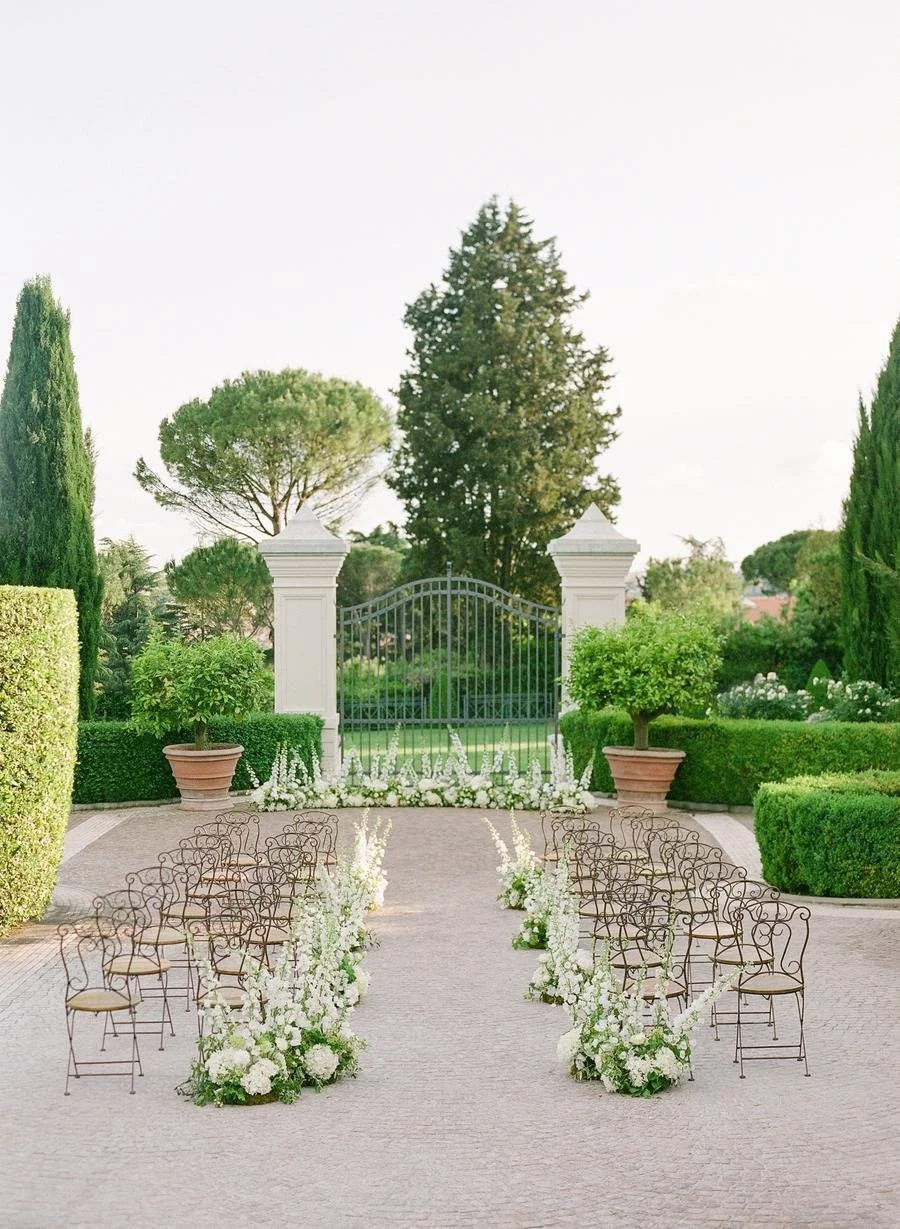 Outdoor wedding ceremony setup with chairs arranged in rows, lined with white flowers, set in a garden with a large gate and trees in the background.