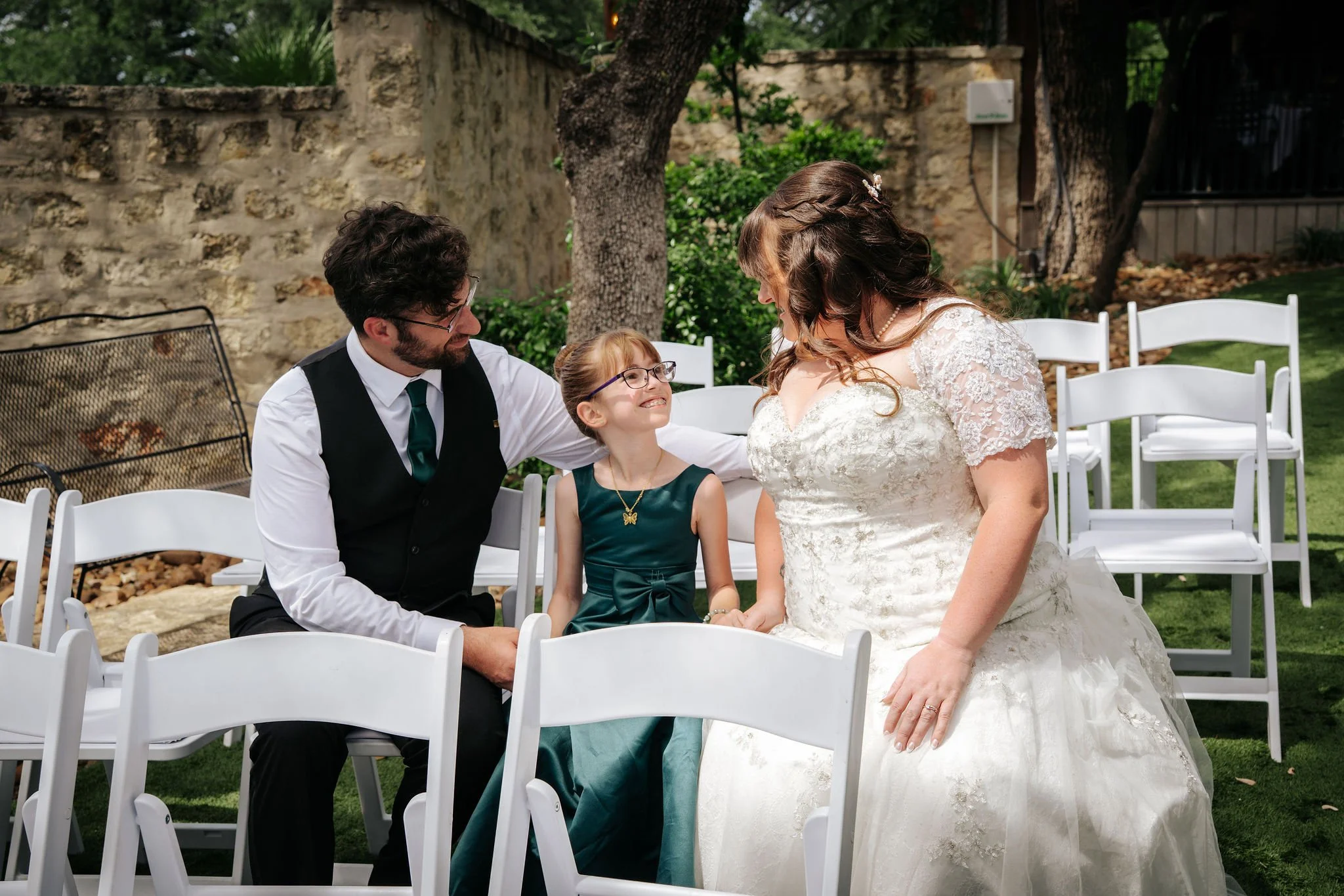 A bride, groom, and a young girl, possibly their daughter, with glasses are sitting outside at their wedding, smiling and looking at each other, surrounded by white folding chairs and trees.