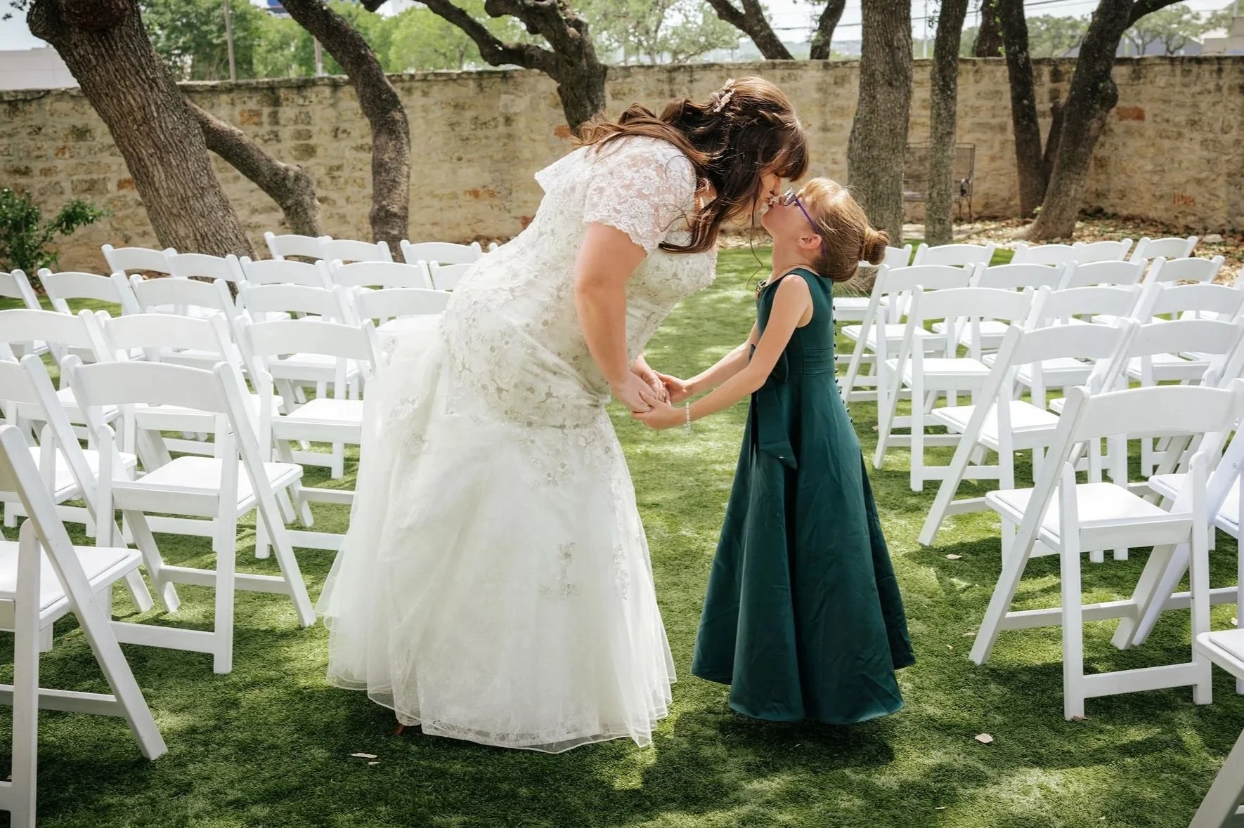 A bride and a young girl, possibly a flower girl, sharing a joyful moment while holding hands and leaning towards each other at an outdoor wedding ceremony. The scene is set on green grass with white chairs arranged in rows, trees, and a stone wall in the background.