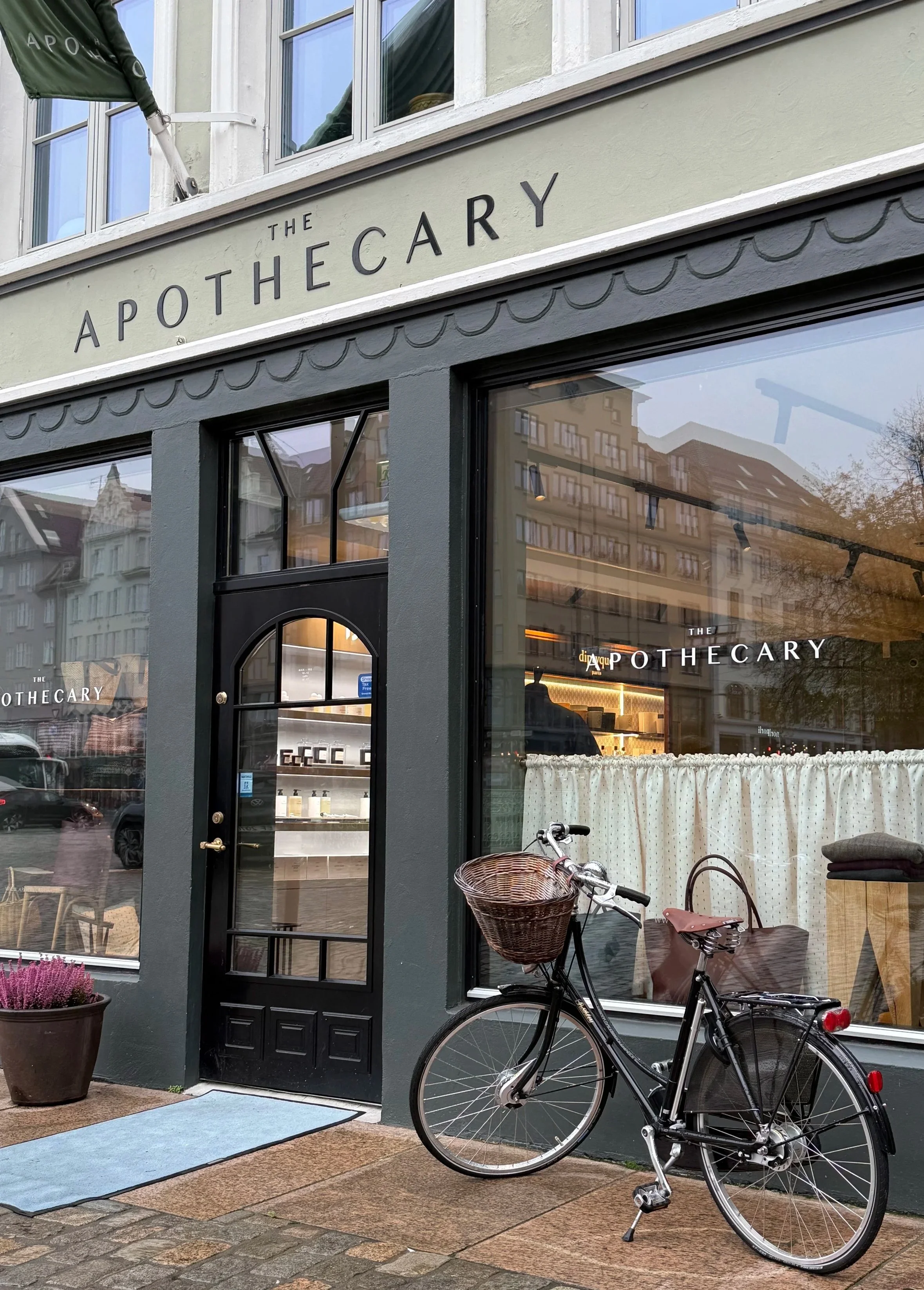 The storefront of 'The Apothecary' with a black door and large glass windows, a bicycle with a basket parked outside, and a potted plant on the sidewalk.