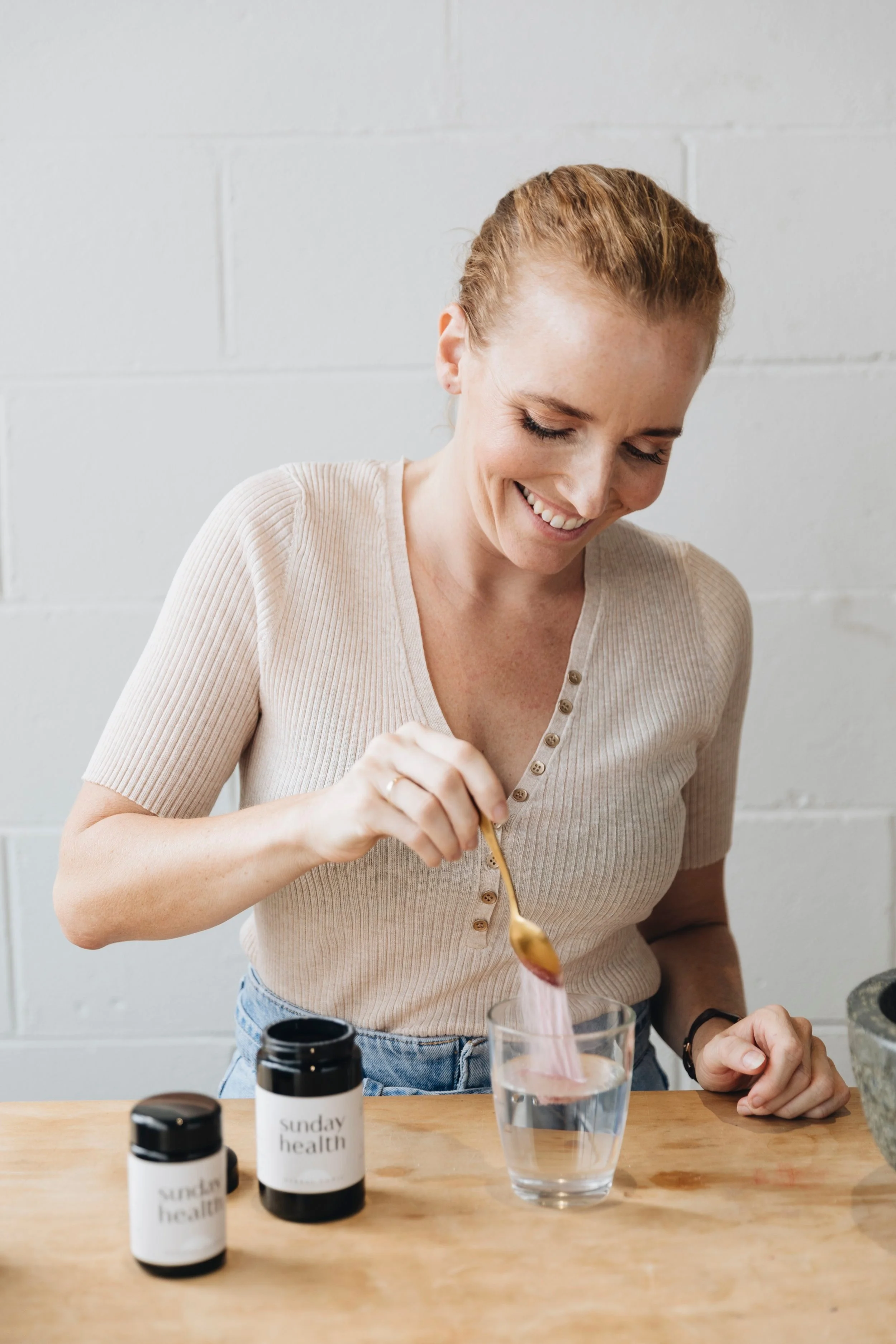 A woman smiling at a glass of water with a pink pill organizer inside, and two bottles labeled 'sunday health' on a wooden table.