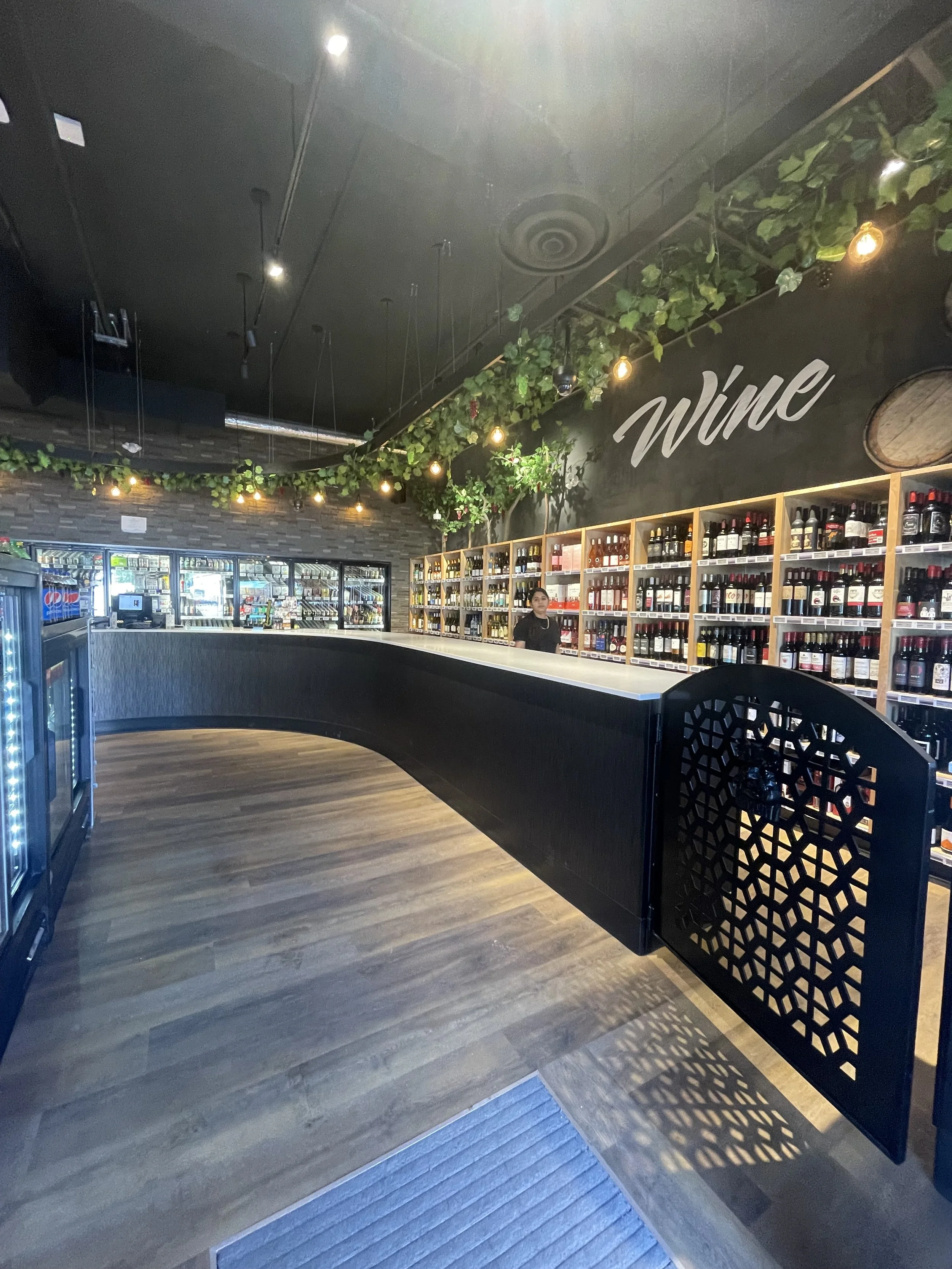 A wine shop with shelves filled with bottles, a black counter, a person behind the counter, and a sign on the wall that says 'Wine'