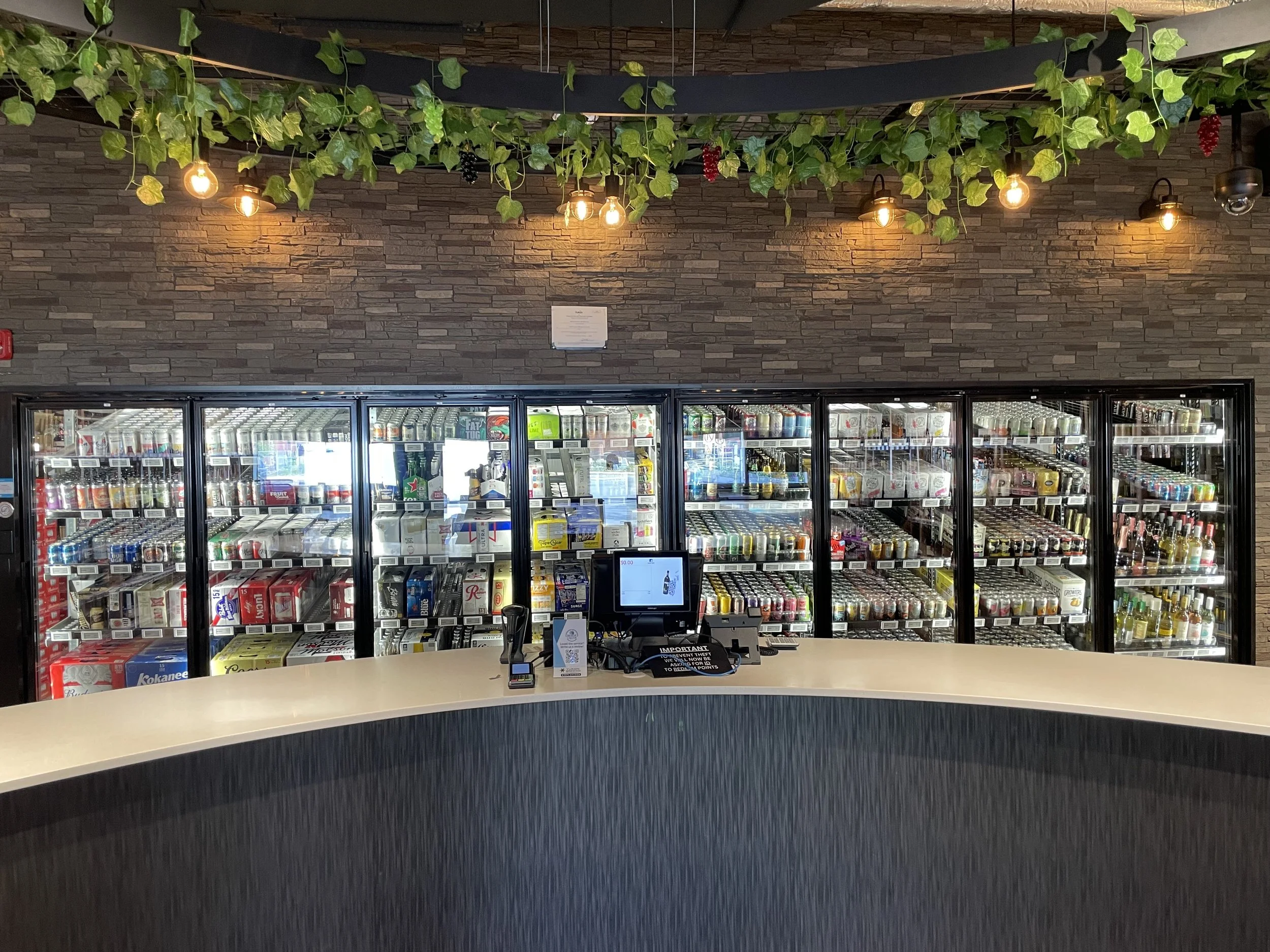 A beverage fridge filled with various bottled and canned drinks behind a curved white counter in a restaurant or bar, with decorative green vine and leaf garland overhead and warm hanging lights.