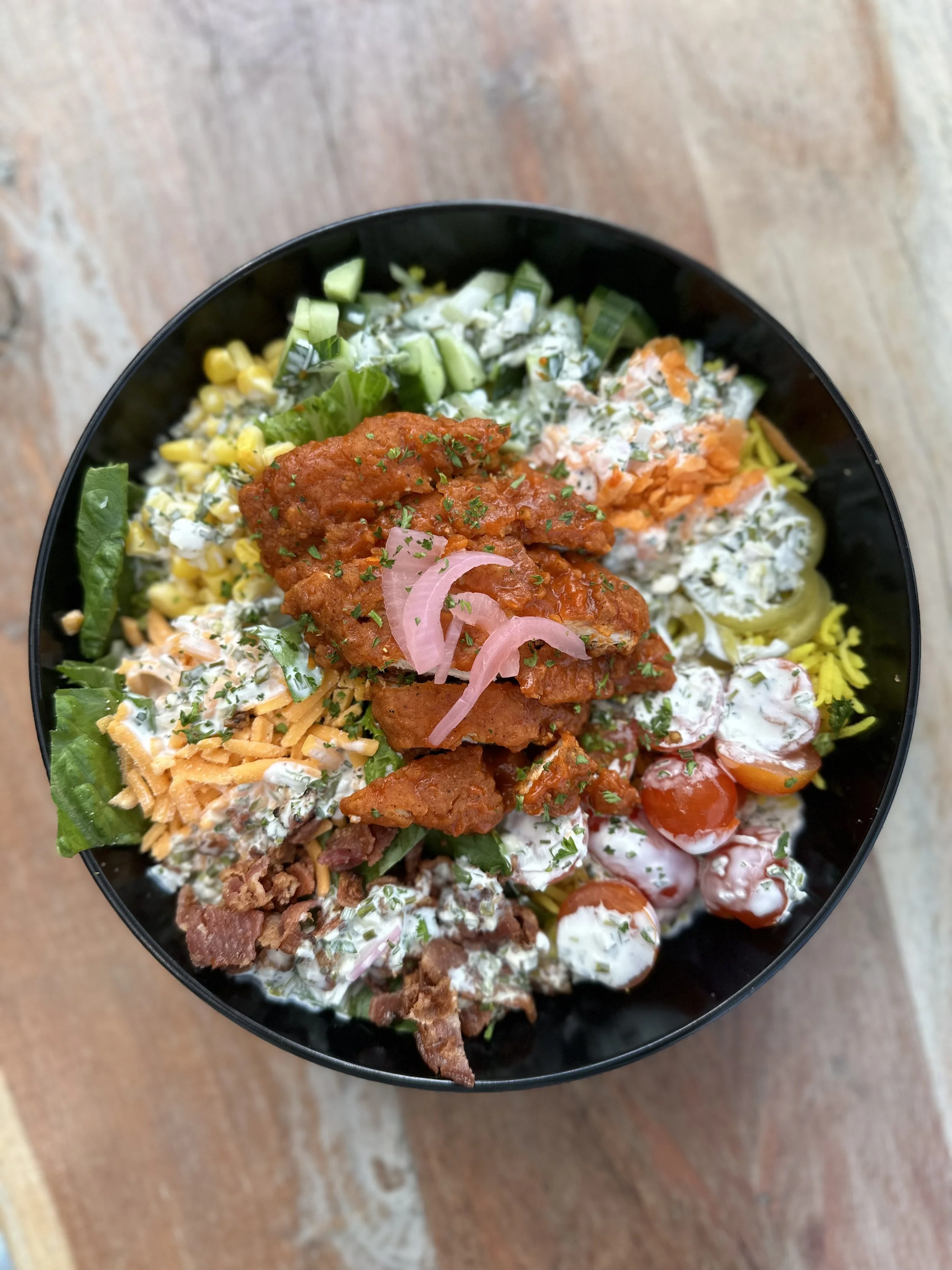 A black bowl filled with a colorful salad containing chopped cucumbers, shredded carrots, cherry tomatoes, corn, lettuce, shredded cheese, crumbled bacon, fried chicken strips topped with pink pickled onions, and various creamy dressings, all on a wooden surface.