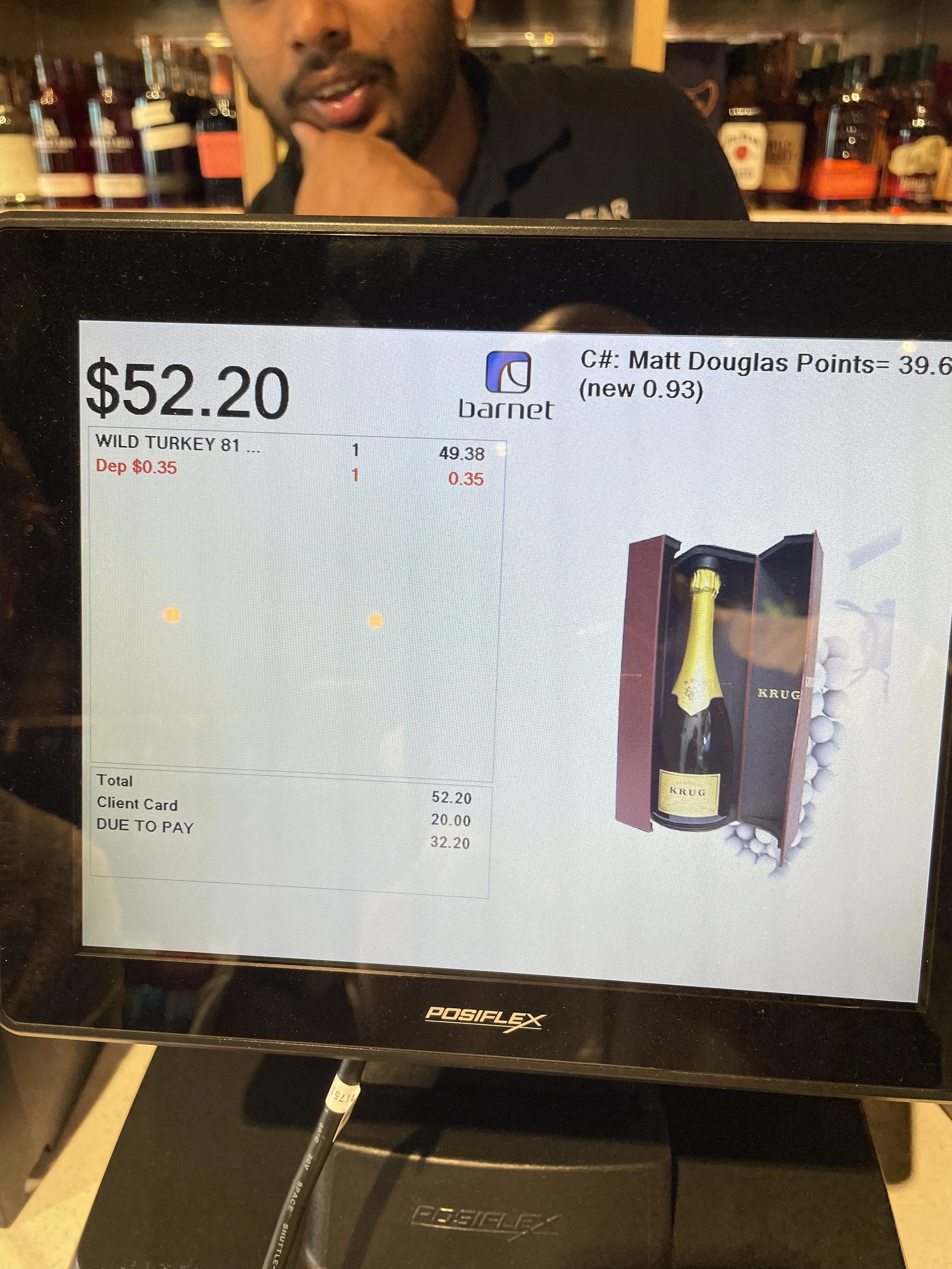 A restaurant receipt on a POS terminal showing a purchase of one bottle of Krug champagne, totaling $52.20, with a background of a bar setting. A man is partially visible behind the screen, appearing to be looking at the camera.