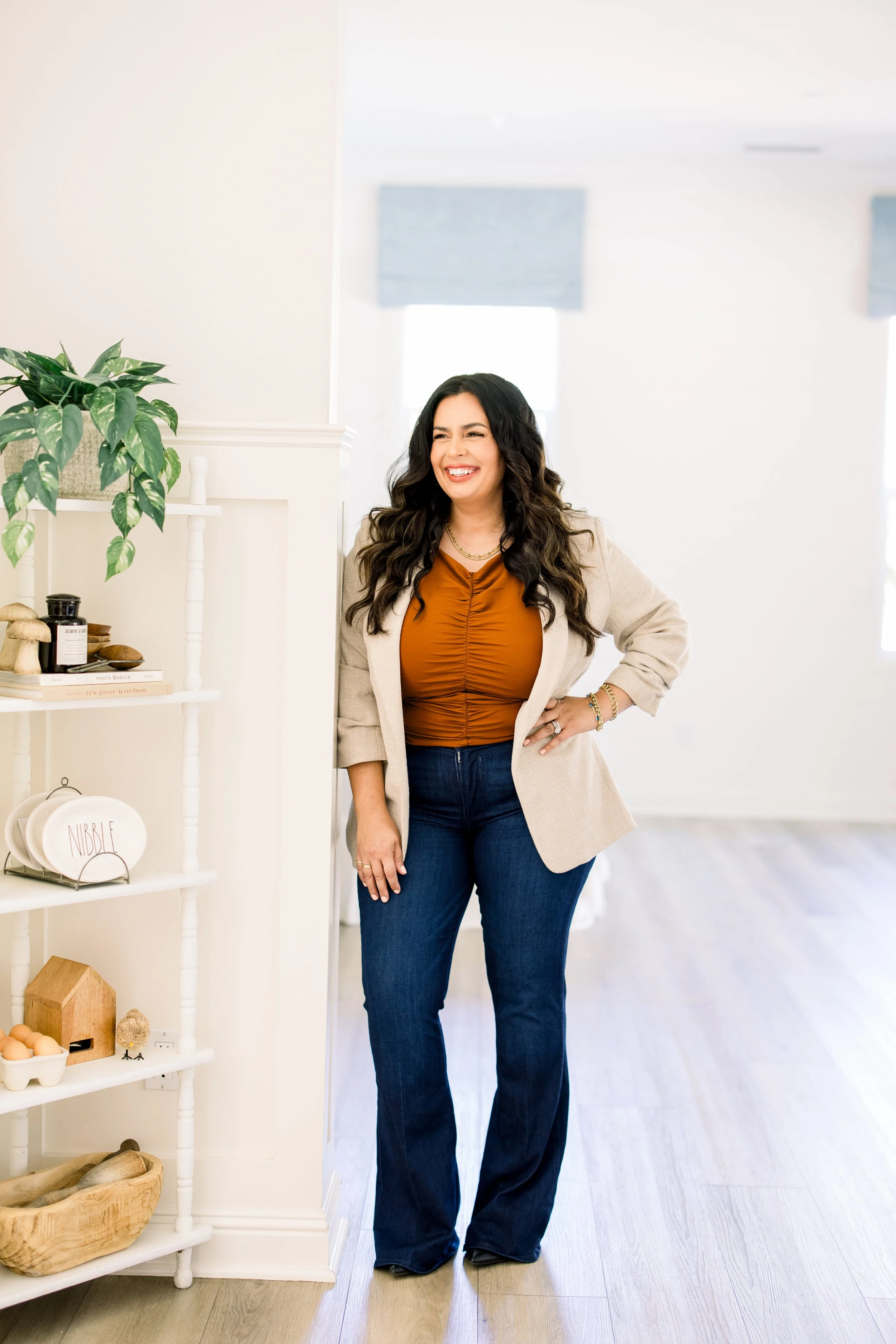 A woman with long dark curly hair, wearing a tan blazer, orange top, and dark blue jeans, is smiling and standing in a bright, modern room next to a white shelf with decorative items and plants.