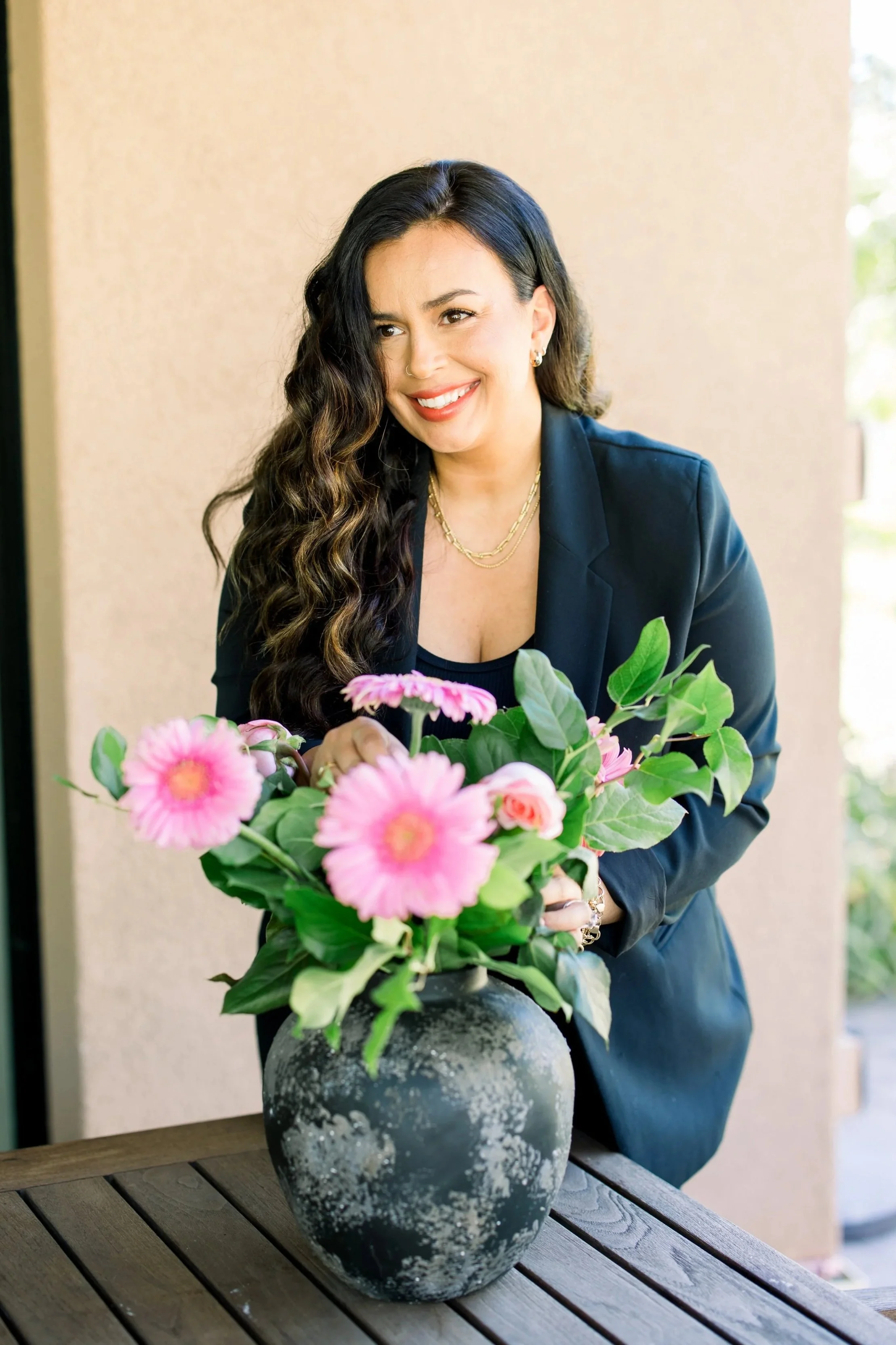 A woman with long wavy dark hair and a bright smile is arranging pink flowers in a black vase with a textured finish on a wooden table. She is wearing a black blazer and gold jewelry, and the background shows a beige wall and some greenery outside.