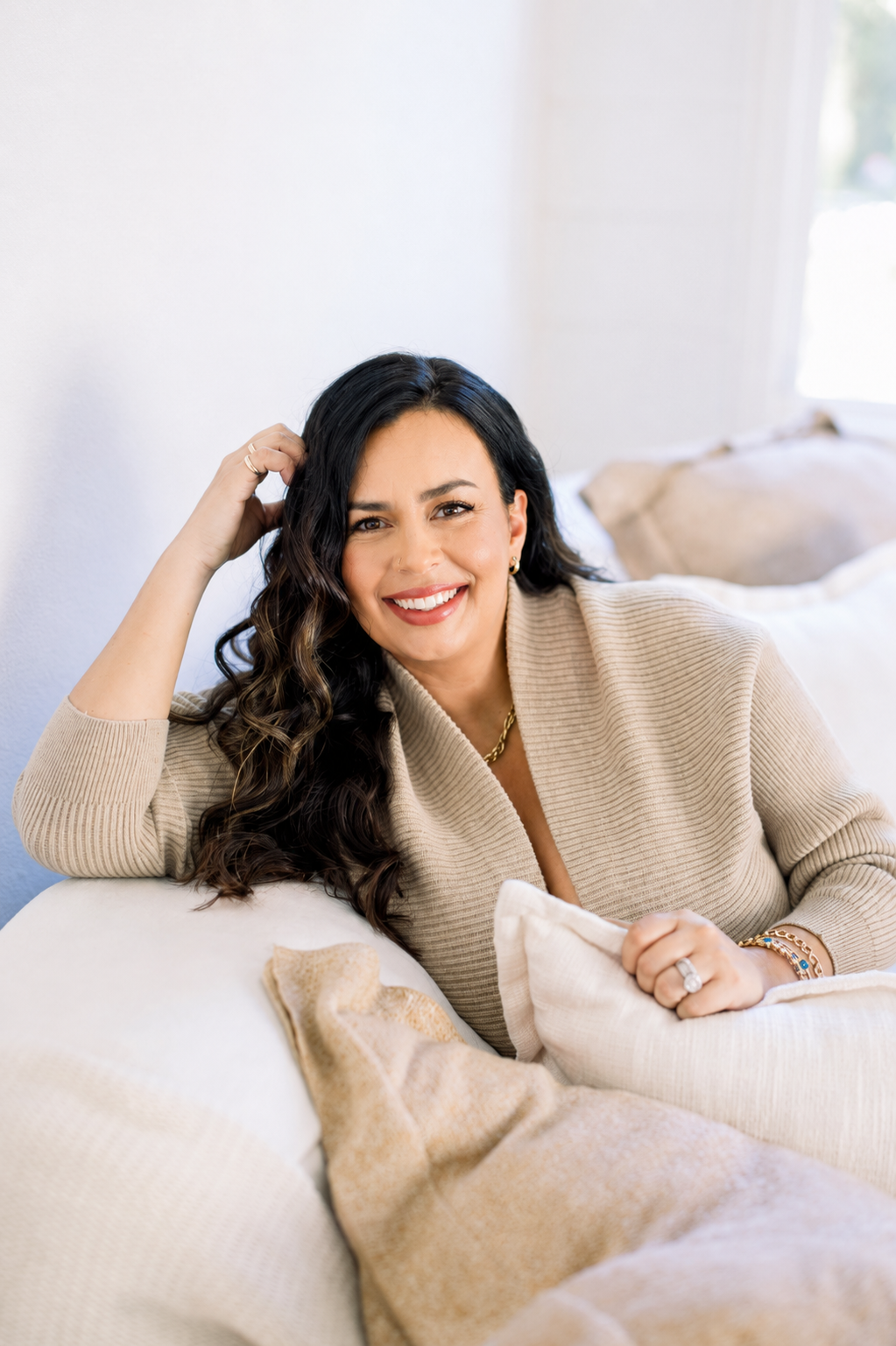 A woman with long, curly, dark hair, smiling, sitting on a beige sofa, wearing a beige sweater, with pillows behind her, in a bright living room.