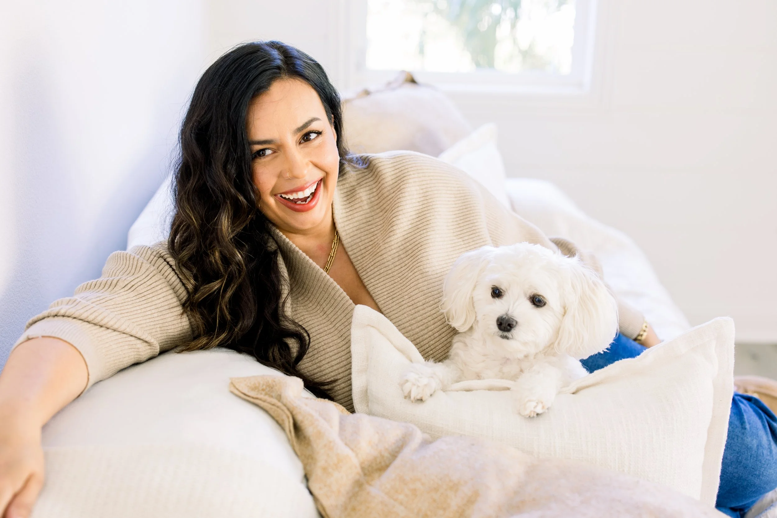 A woman with long dark hair and a light beige sweater, smiling and lying on a white couch, with a small white dog lying beside her.