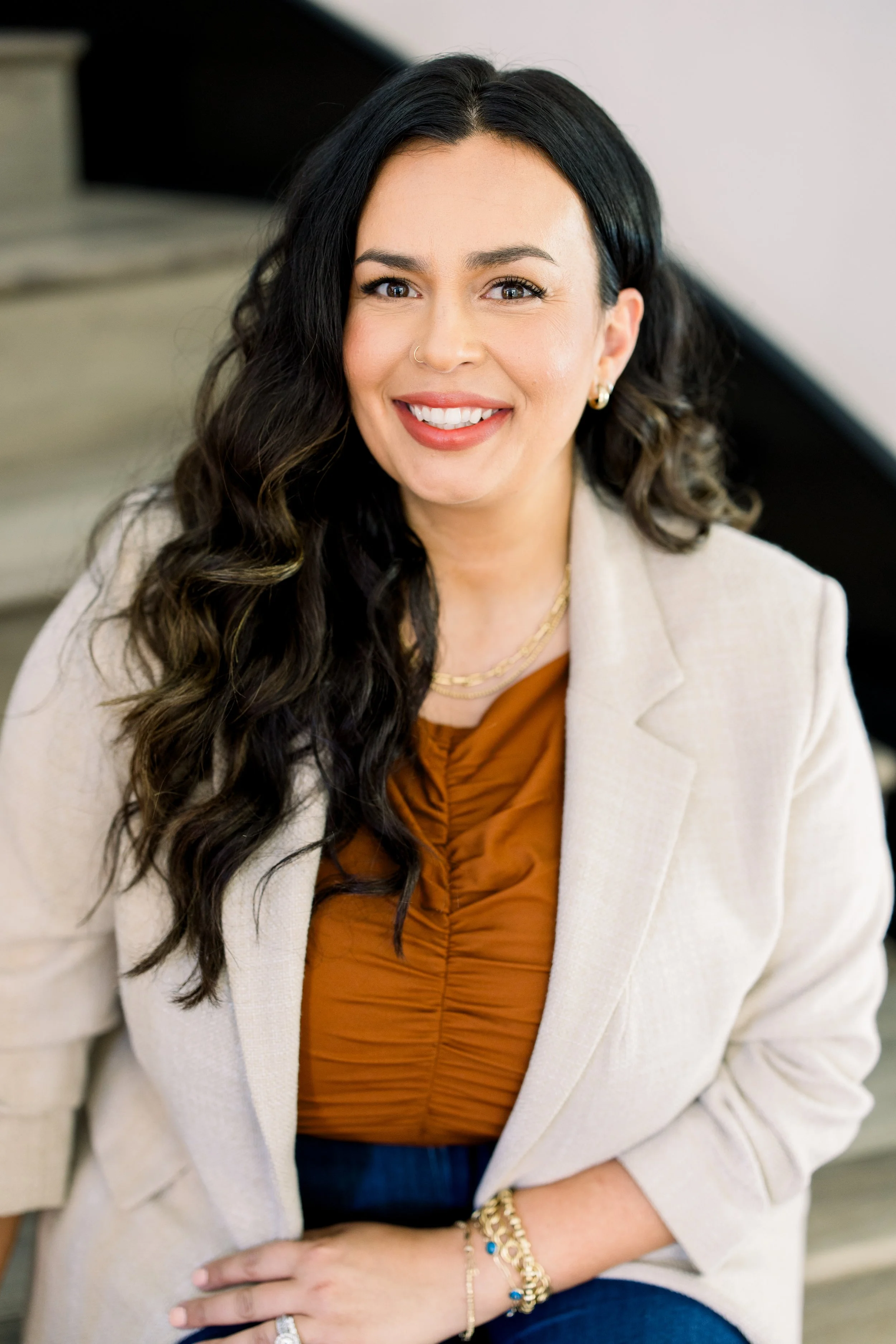 A woman with long dark curled hair, wearing a beige blazer over an orange top, smiling, with jewelry including earrings, a necklace, bracelets, and a ring.