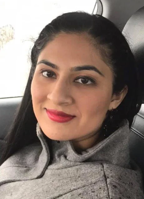 A young woman with long dark hair and red lipstick sitting in a car, wearing a gray hoodie.
