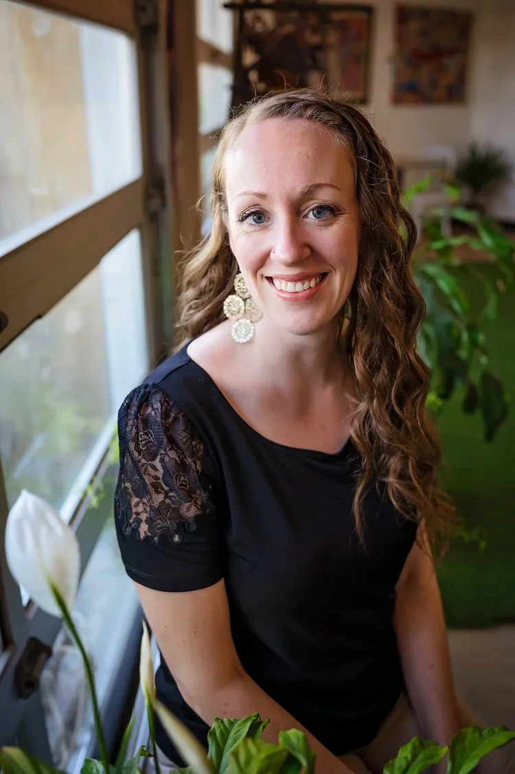 A smiling woman with long curly hair wearing a black top with lace sleeves, sitting indoors near a large window with greenery in the background.