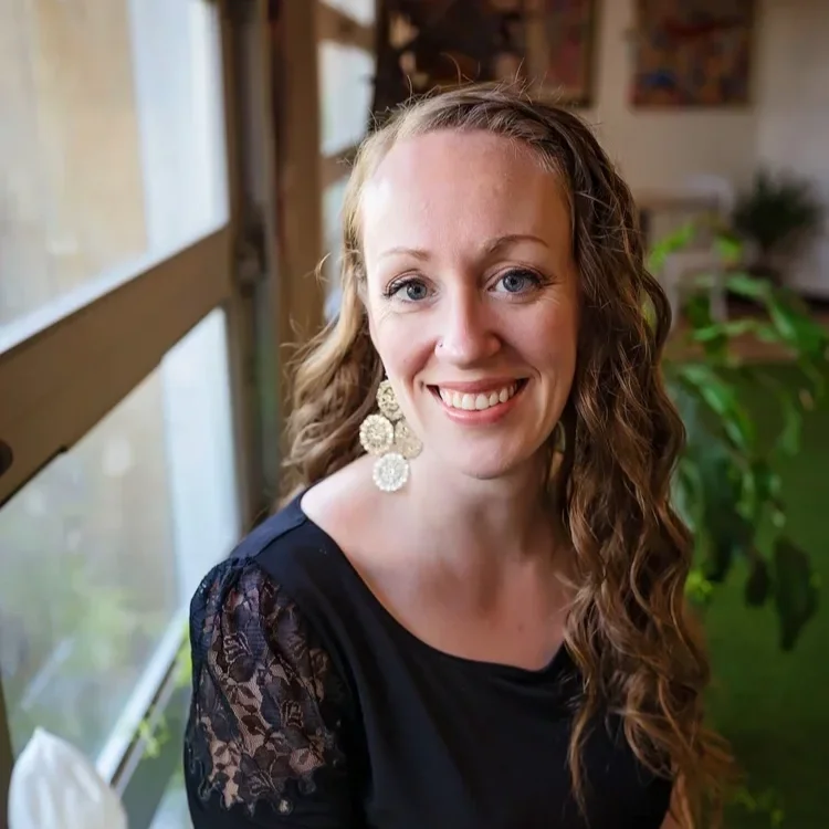A woman with curly hair smiling, wearing large floral earrings and a black top with lace detail on the sleeves, seated indoors near a window with plants in the background.