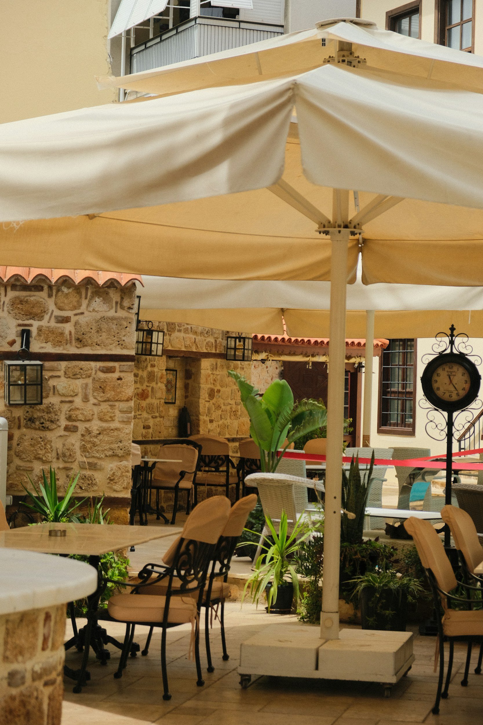 An outdoor patio area with large beige umbrellas, stone walls, potted plants, and various chairs and tables, some with cushions.