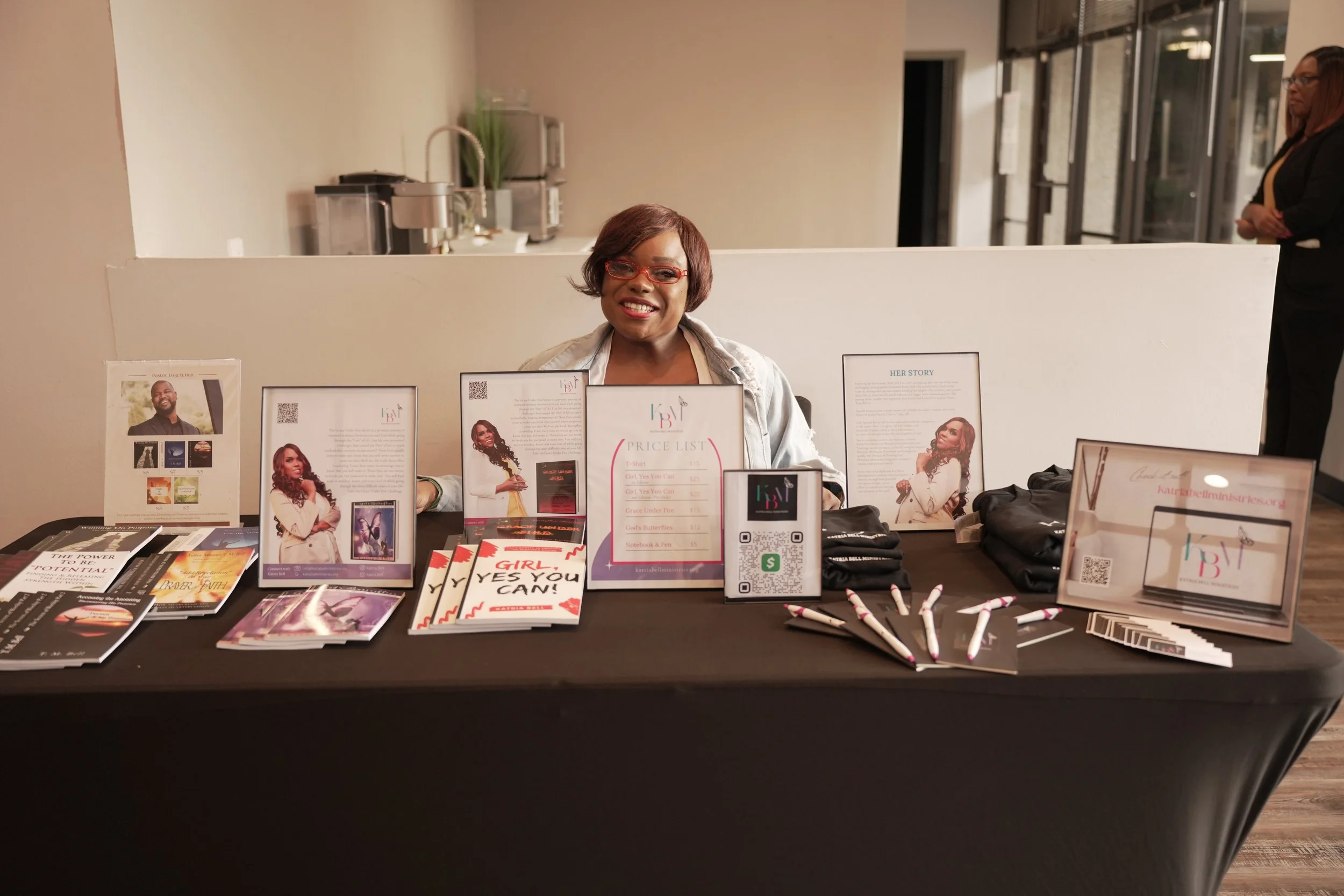 A smiling woman sitting at a table with various promotional materials, books, pens, and framed photos displayed in front of her, with two other women standing in the background.