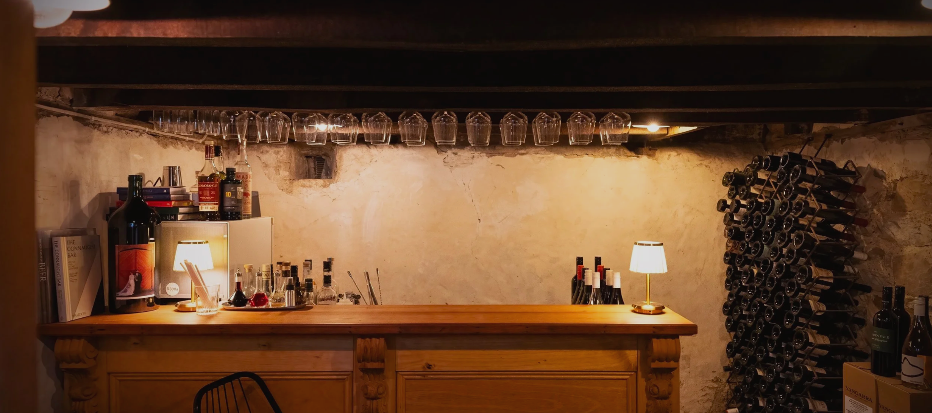 A cozy bar with a wooden counter, glassware hanging upside down above, wine bottles stored on a rack, and small lamps providing warm lighting.
