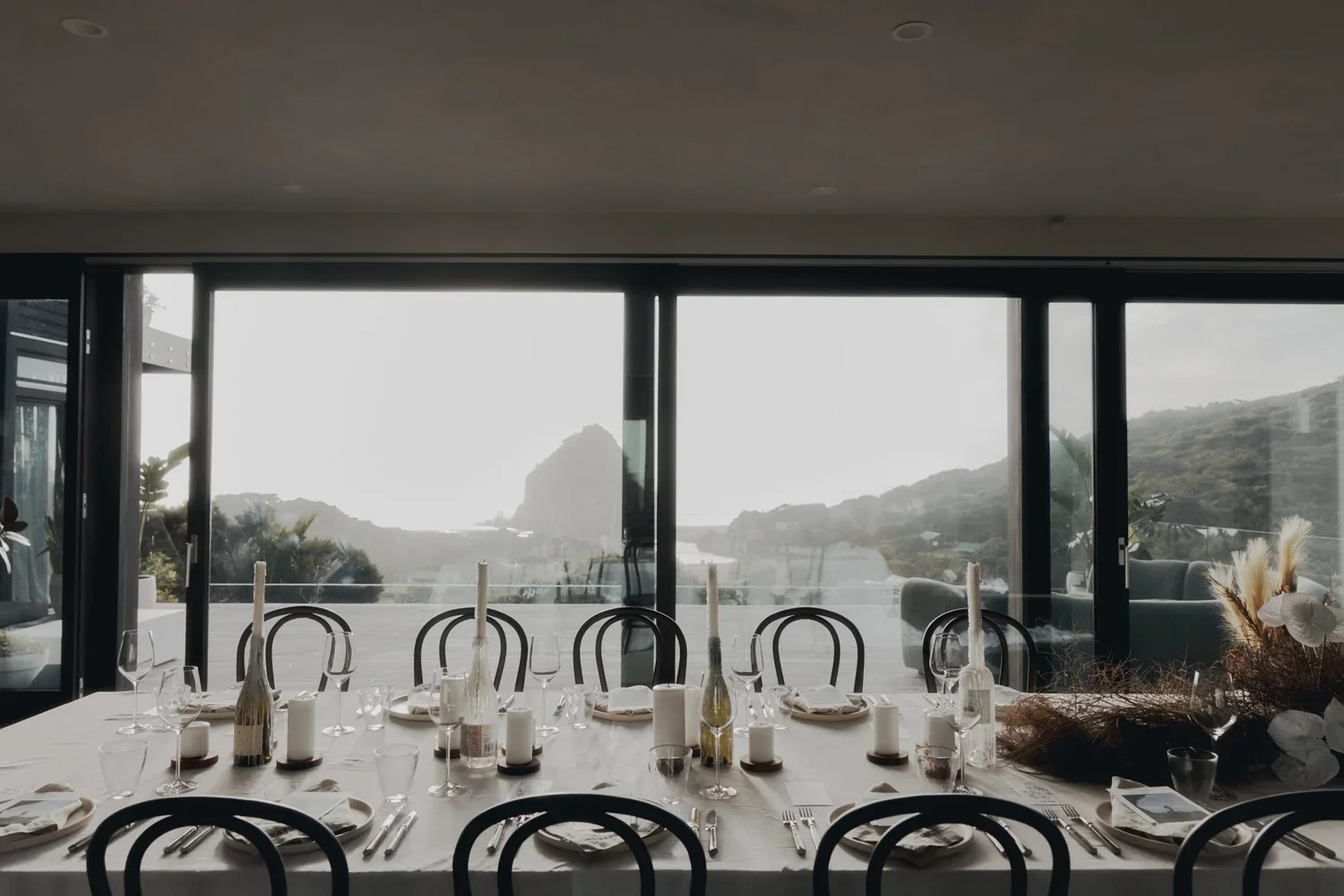 Elegant dining table set with candles, wine glasses, and tableware in front of large glass windows overlooking a scenic mountain and water landscape.