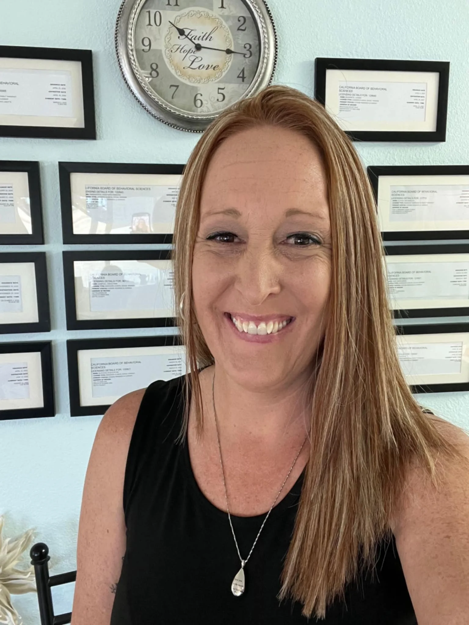 A smiling woman with long reddish-brown hair wearing a black sleeveless top and a silver necklace stands in front of a wall with framed certificates and a decorative clock with the words 'Faith Hope Love' on the face.
