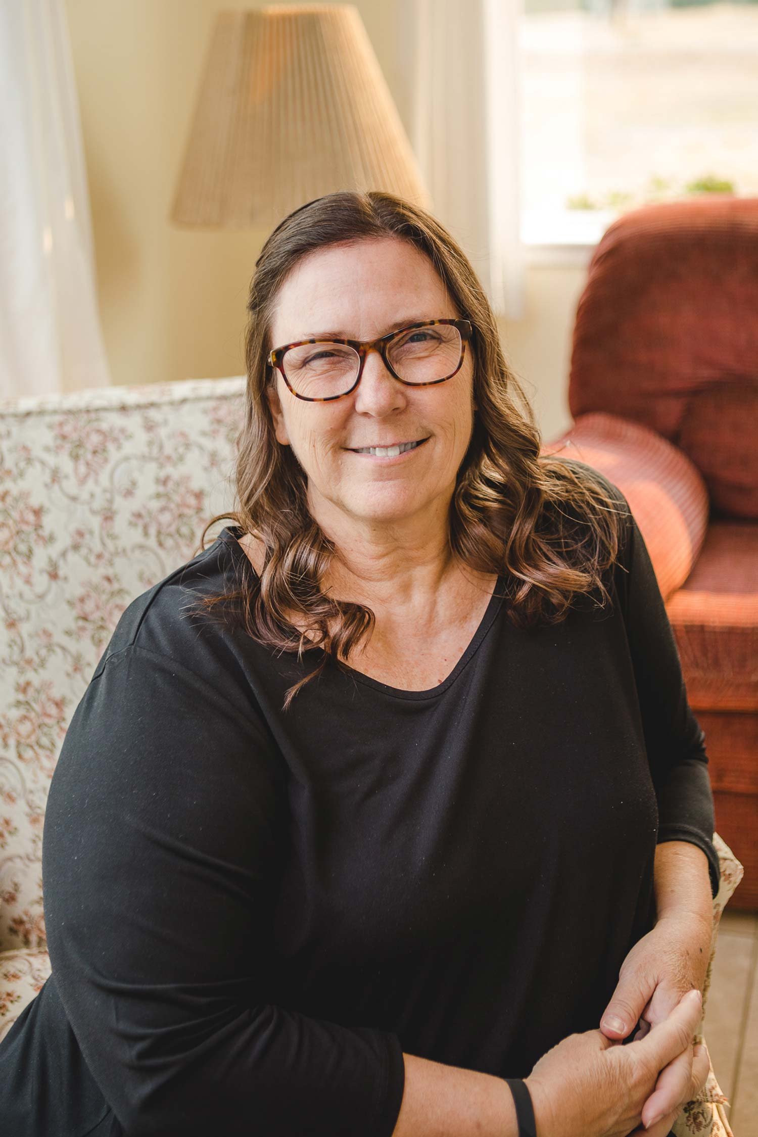 A woman with brown wavy hair, wearing glasses and a black shirt, sitting on a floral armchair in a cozy room with warm lighting, a lamp, and a window in the background.