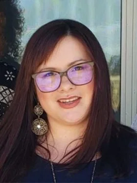 A woman with long dark hair, wearing purple glasses, large dangling earrings, and a dark top, smiling in front of a window with a scenic view.