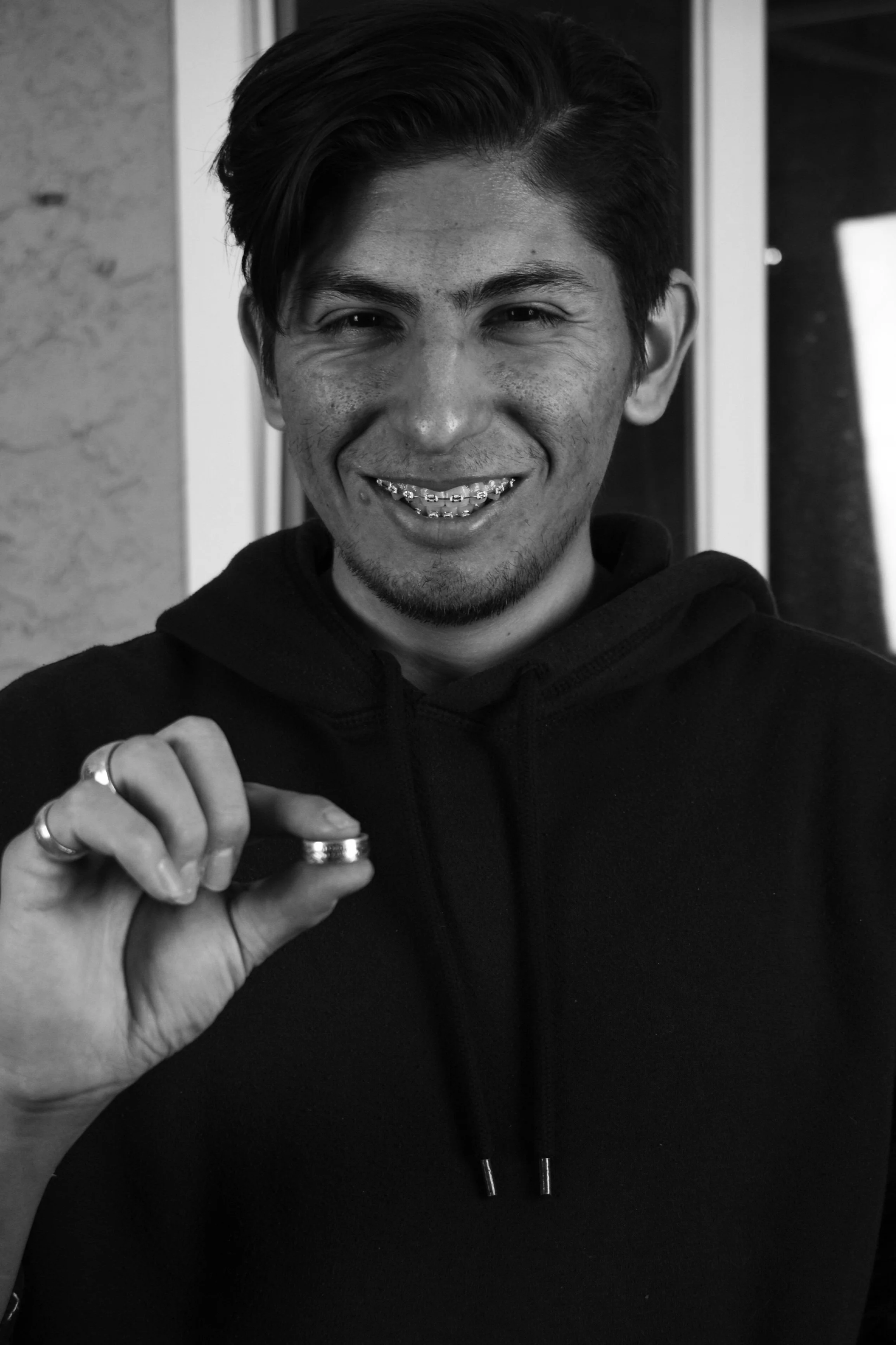 A young man with dark hair, smiling, showing braces, holding a small object in his hand. He is wearing a black hoodie and rings on his fingers.