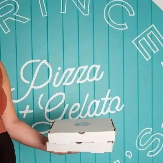 Person holding two stacked pizza boxes in front of a teal wall with white lettering reading "Eat, Drink, Be Happy, Pizza + Gelato".