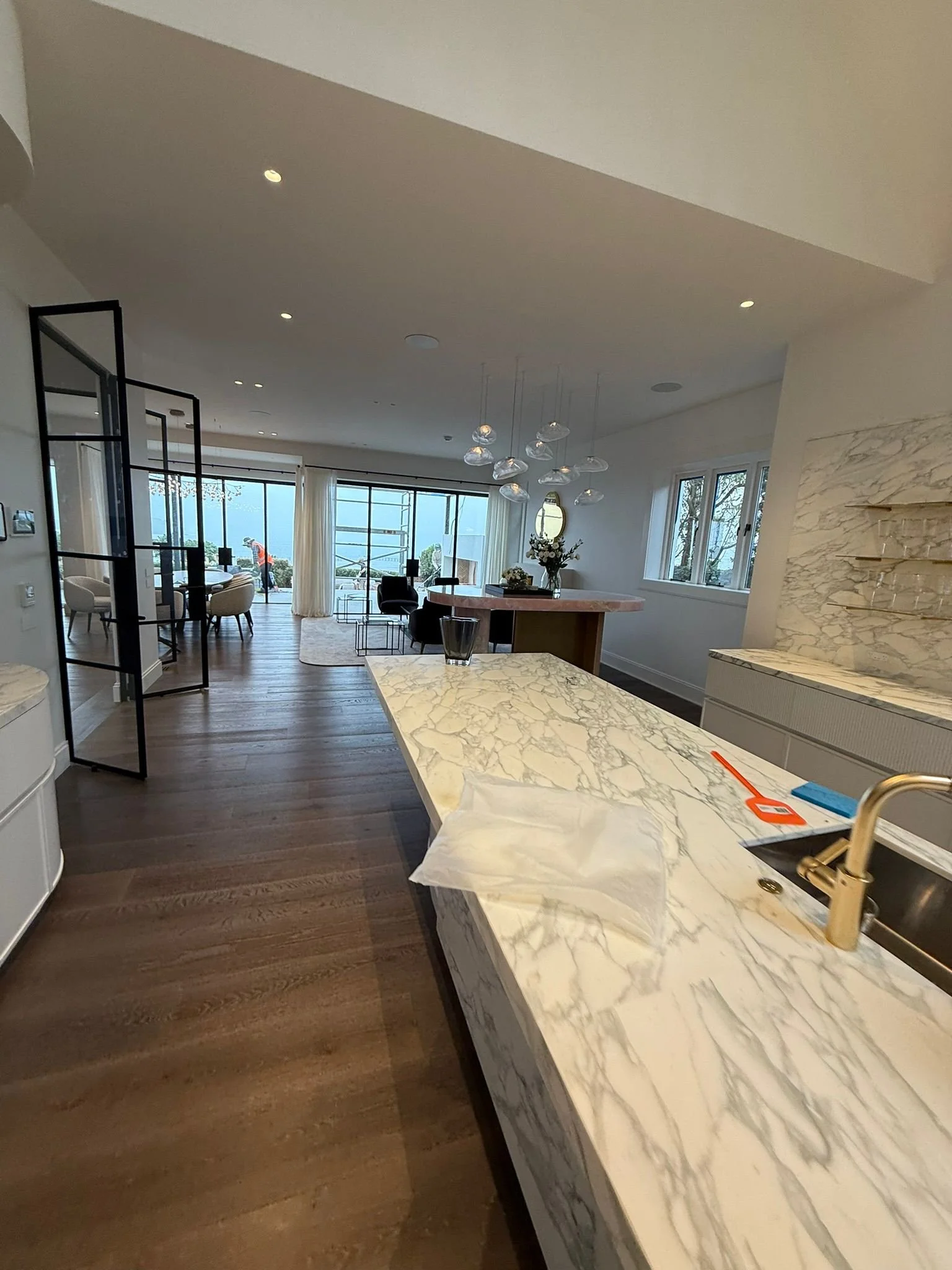 Modern kitchen with marble countertops, open living area with dining and seating, large windows with curtain, wooden flooring, and contemporary lighting fixtures.