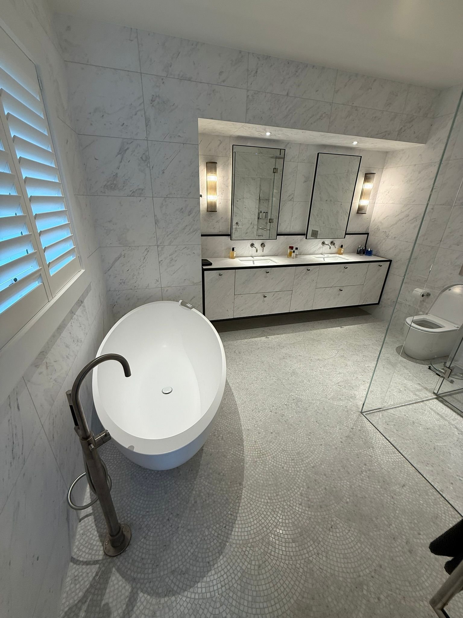 A modern bathroom with a freestanding bathtub, a large double sink vanity with two mirrors, a glass-enclosed shower, and a toilet. The walls and floors are covered in white marble tile.