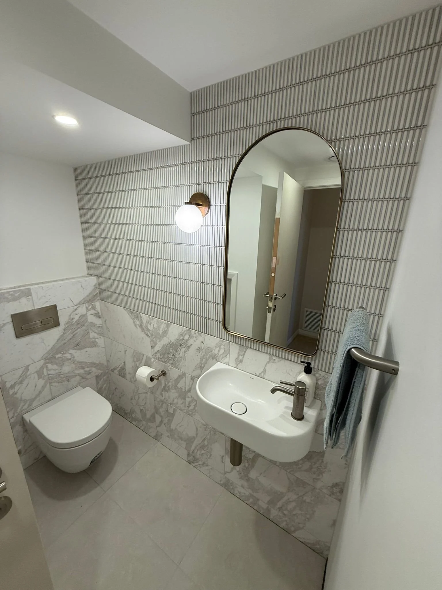 Modern bathroom with small white sink, wall-mounted toilet, large oval mirror, and textured tiles on the wall. There is a wall-mounted light fixture and a towel hanging on a rail.
