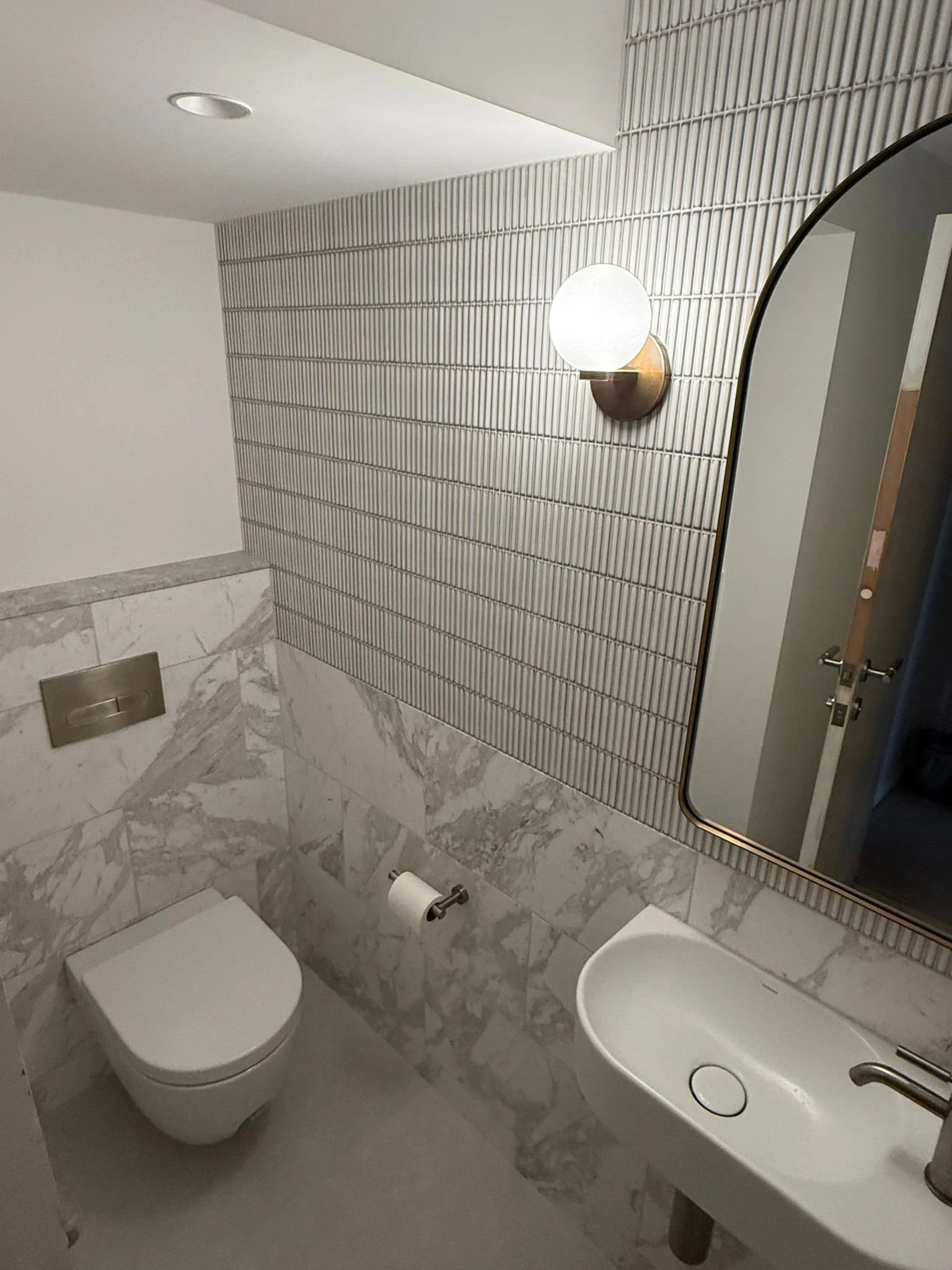 Small modern bathroom with marble half-wall, a white toilet, a small white sink, mirror, and wall-mounted light fixture.