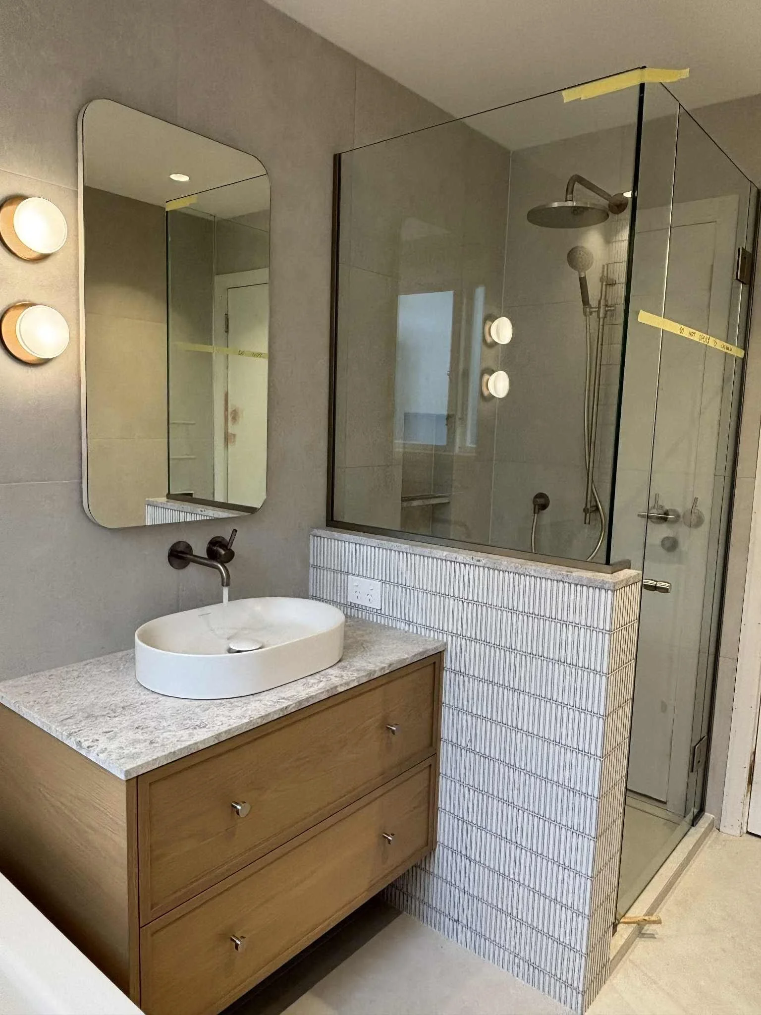 Modern bathroom with a wooden vanity with two drawers, circular sink, wall-mounted black faucet, mirrored medicine cabinet, and a glass-enclosed shower with a rainfall showerhead.