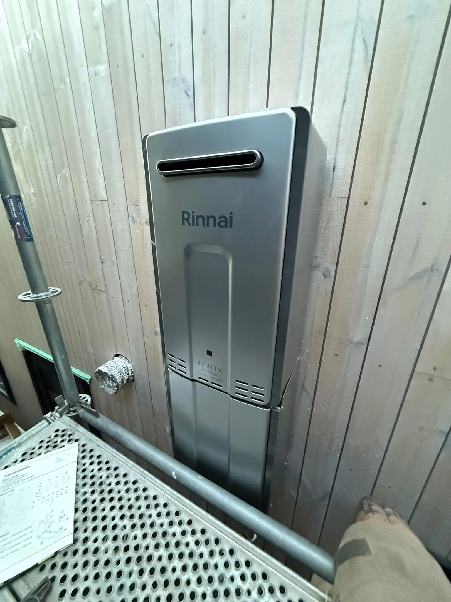 A metal Rinnai exterior tankless water heater mounted on a wooden wall inside a building.
