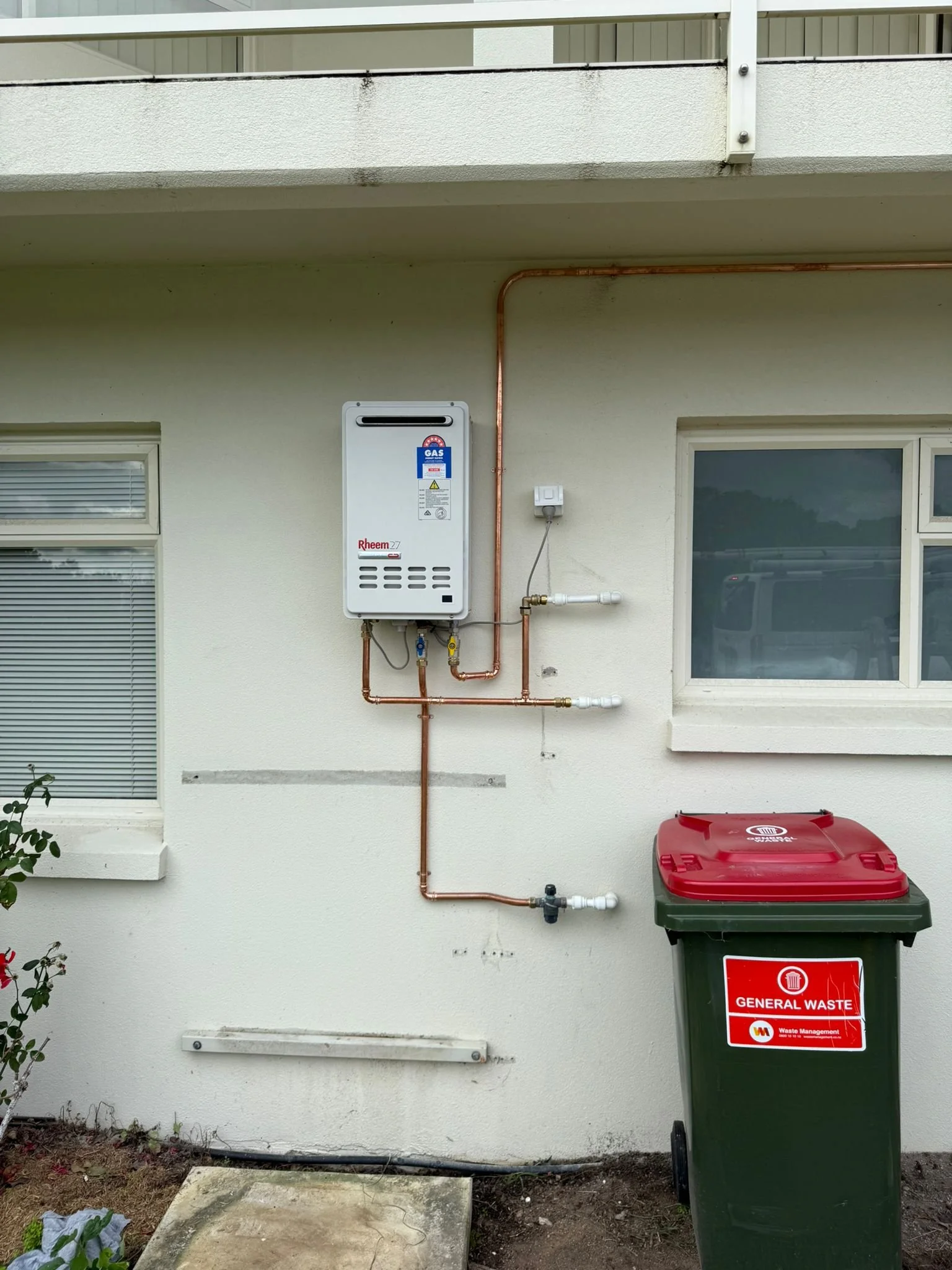 Exterior wall with a gas water heater, copper pipes, a window, and a green waste bin with a red lid labeled 'General Waste'.