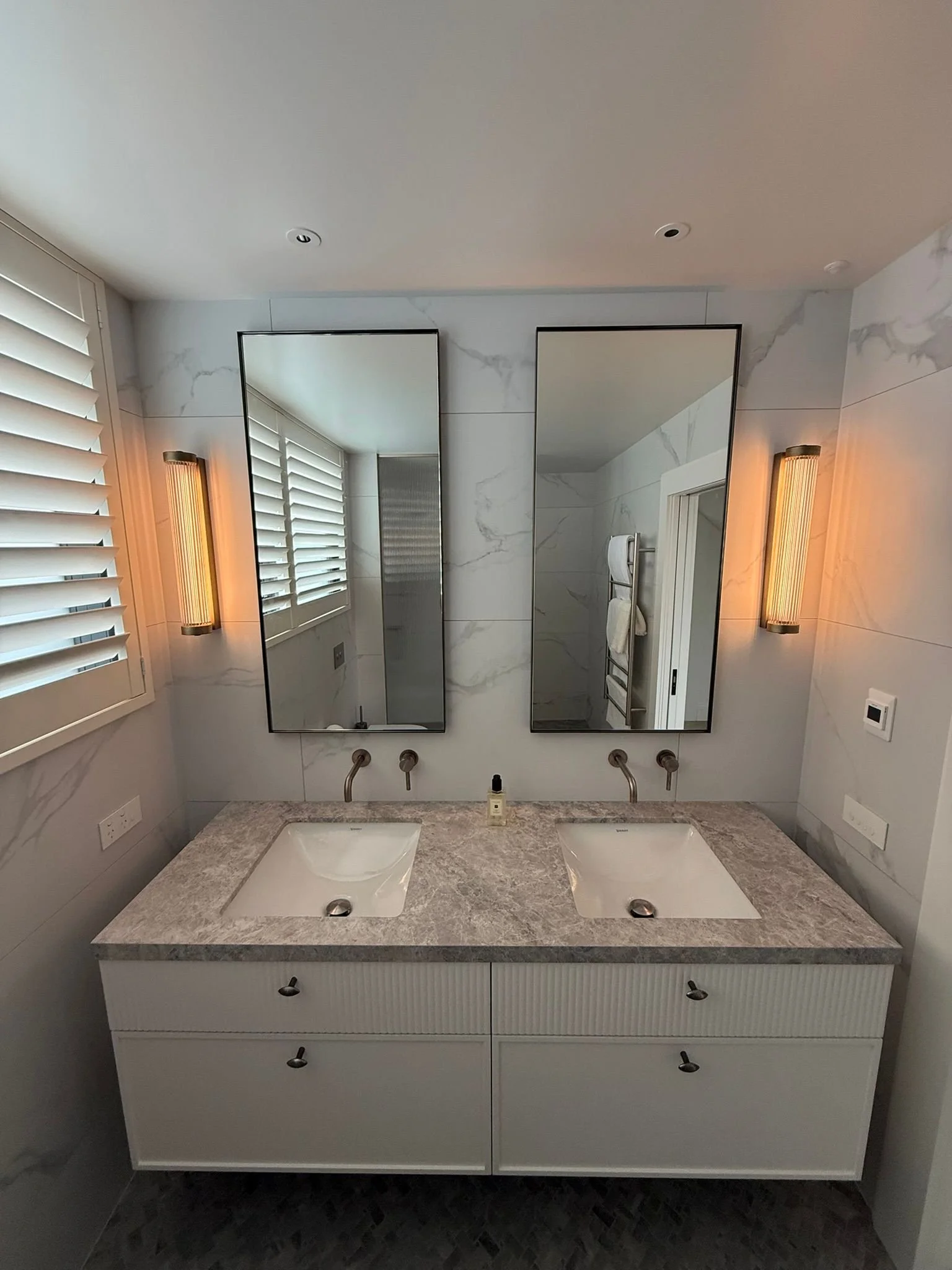 Modern bathroom vanity with two rectangular mirrors, double sinks with stone countertop, wall-mounted faucet, large window with plantation shutters, and wall sconces on either side of the mirrors.