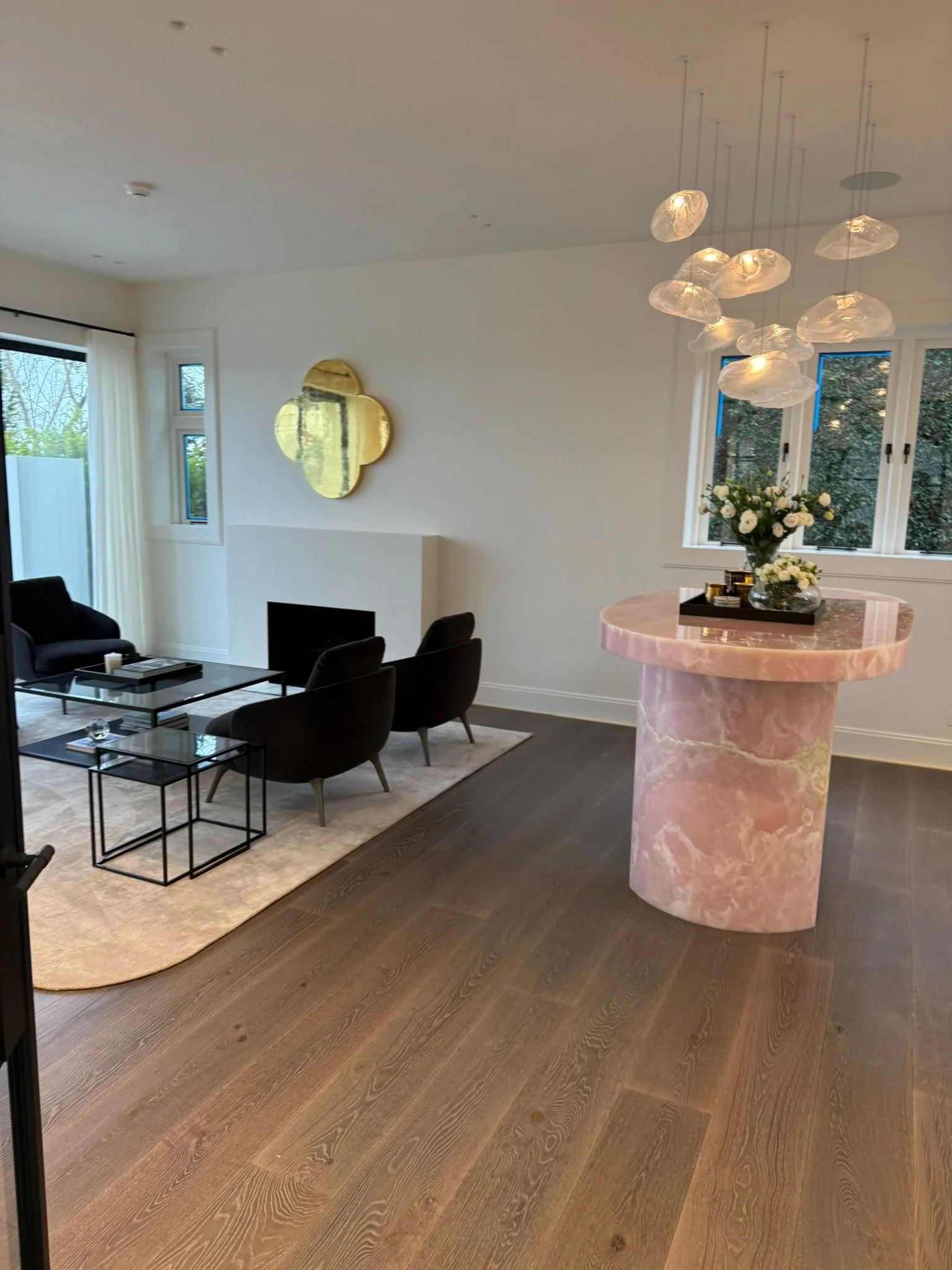Modern living room with black chairs, glass tables, a fireplace, a gold decorative mirror, a pink marble side table with a flower arrangement, and a contemporary chandelier with glass cloud-like shades.