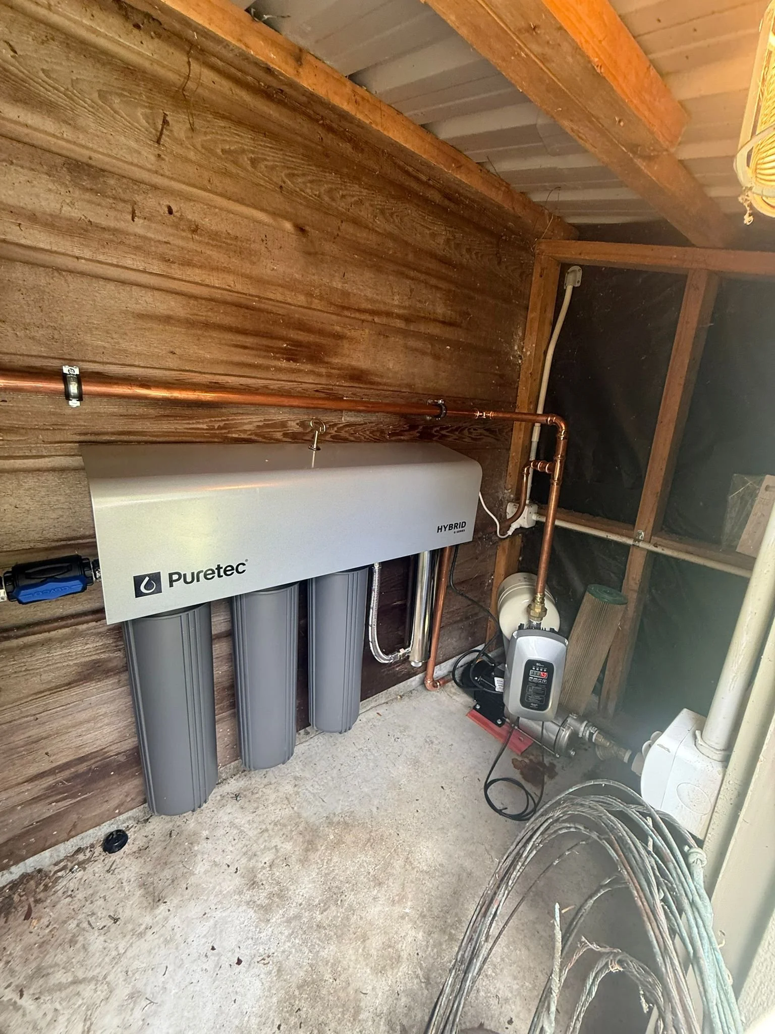 A water filtration system installed in a wooden shed, with copper pipes, a Puretec Hybrid filter, and related plumbing components.