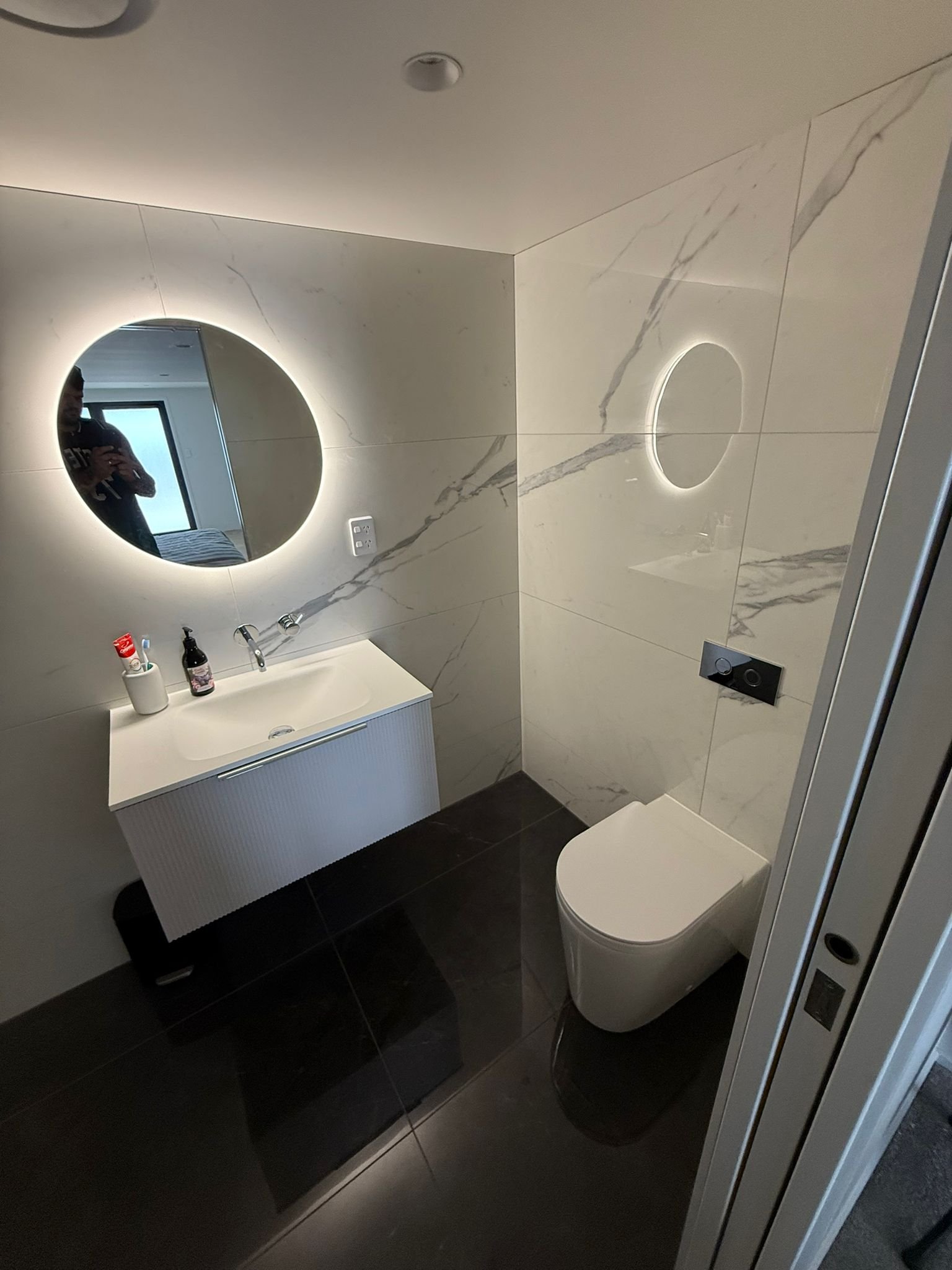 Modern bathroom with a circular backlit mirror, a small white vanity with toiletries, a wall-mounted toilet, and marble wall tiles.