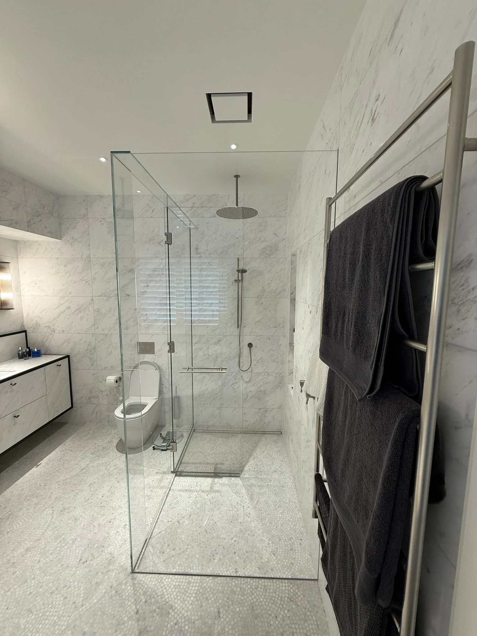 Modern bathroom with a walk-in glass shower, black towels on a metallic towel rack, a white toilet, a white marble vanity, and a window with blinds.