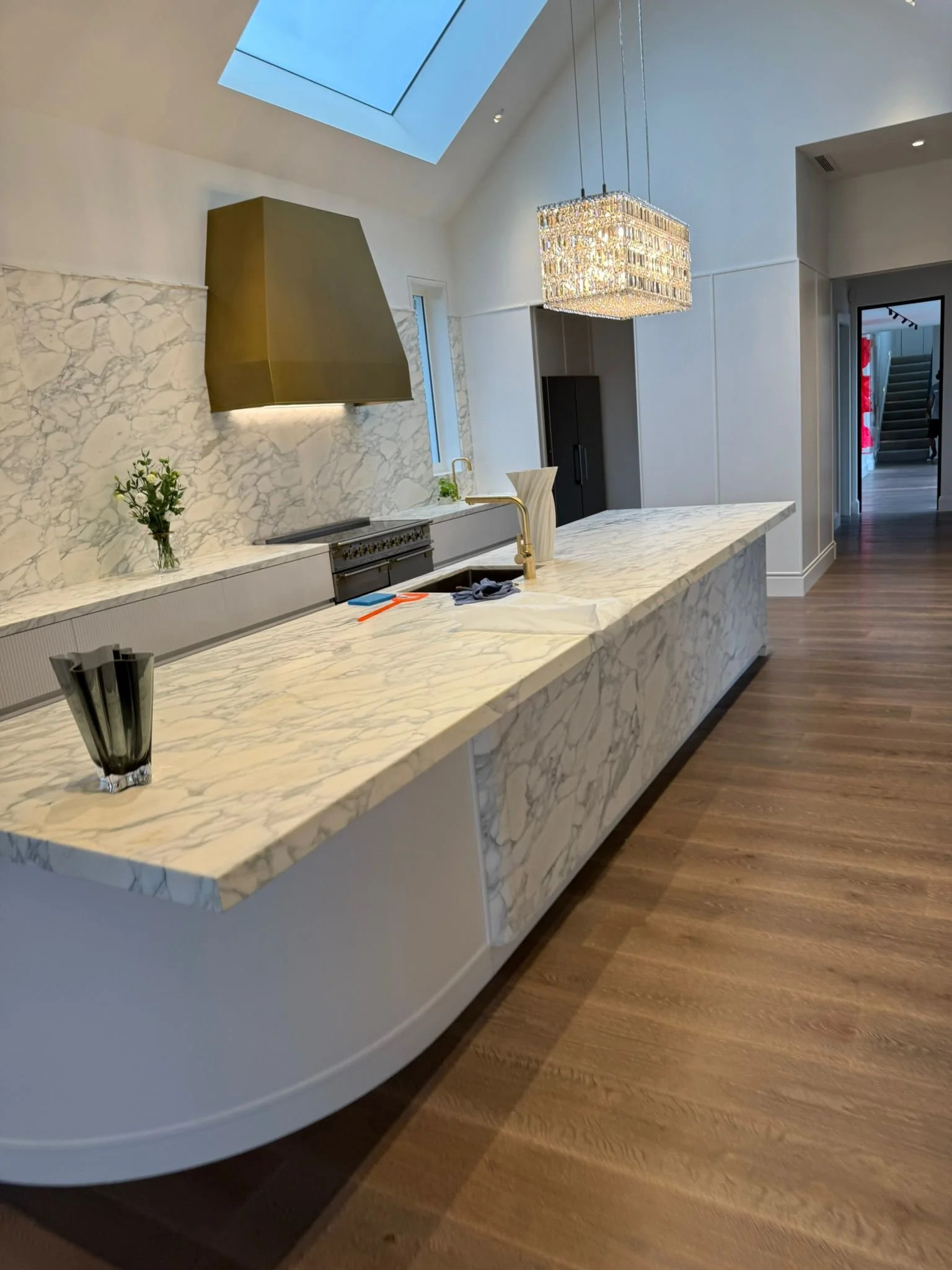 Modern kitchen with marble countertops and backsplash, gold range hood, and elegant chandelier, open to a hallway with wooden floors.